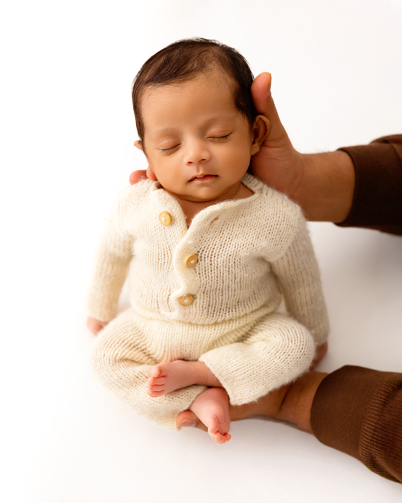 Newborn baby in cream knit romper with eyes closed, gently supported by adult hands on white background.