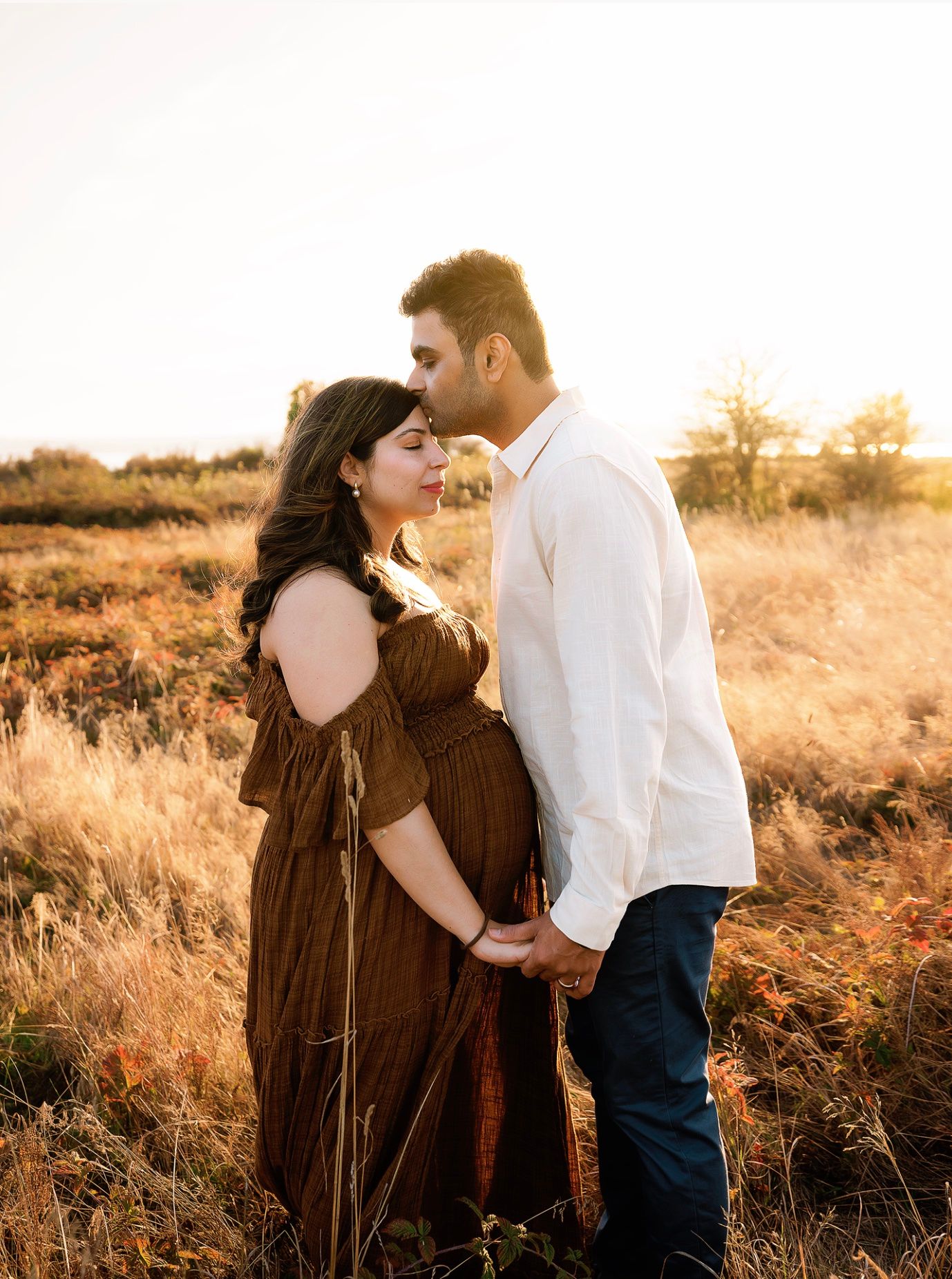 Couple sharing a tender moment in golden field at sunset, pregnant woman in brown dress held by man in white shirt.