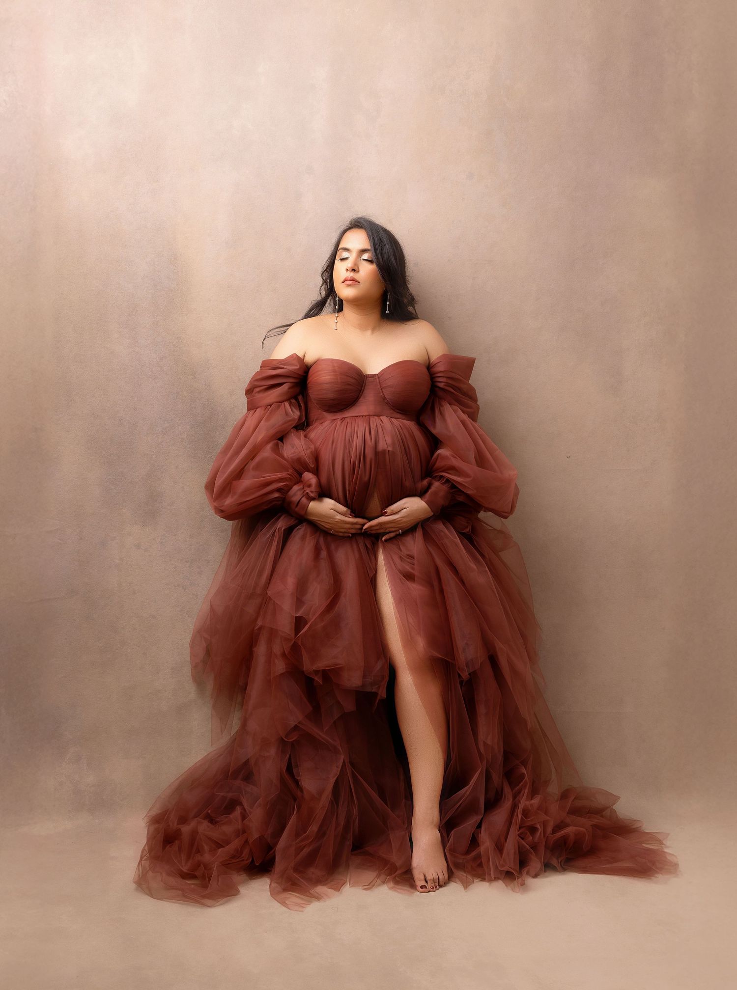 Pregnant woman in elegant rust tulle off-shoulder maternity gown poses gracefully against neutral studio backdrop.