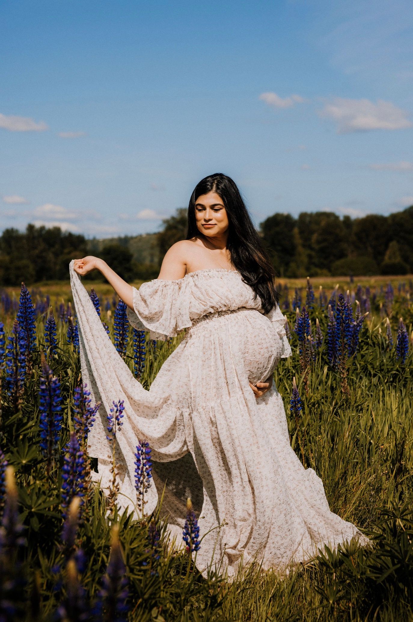 A flowing white lace dress twirls gracefully among purple lupine wildflowers in a sunlit meadow during golden hour.