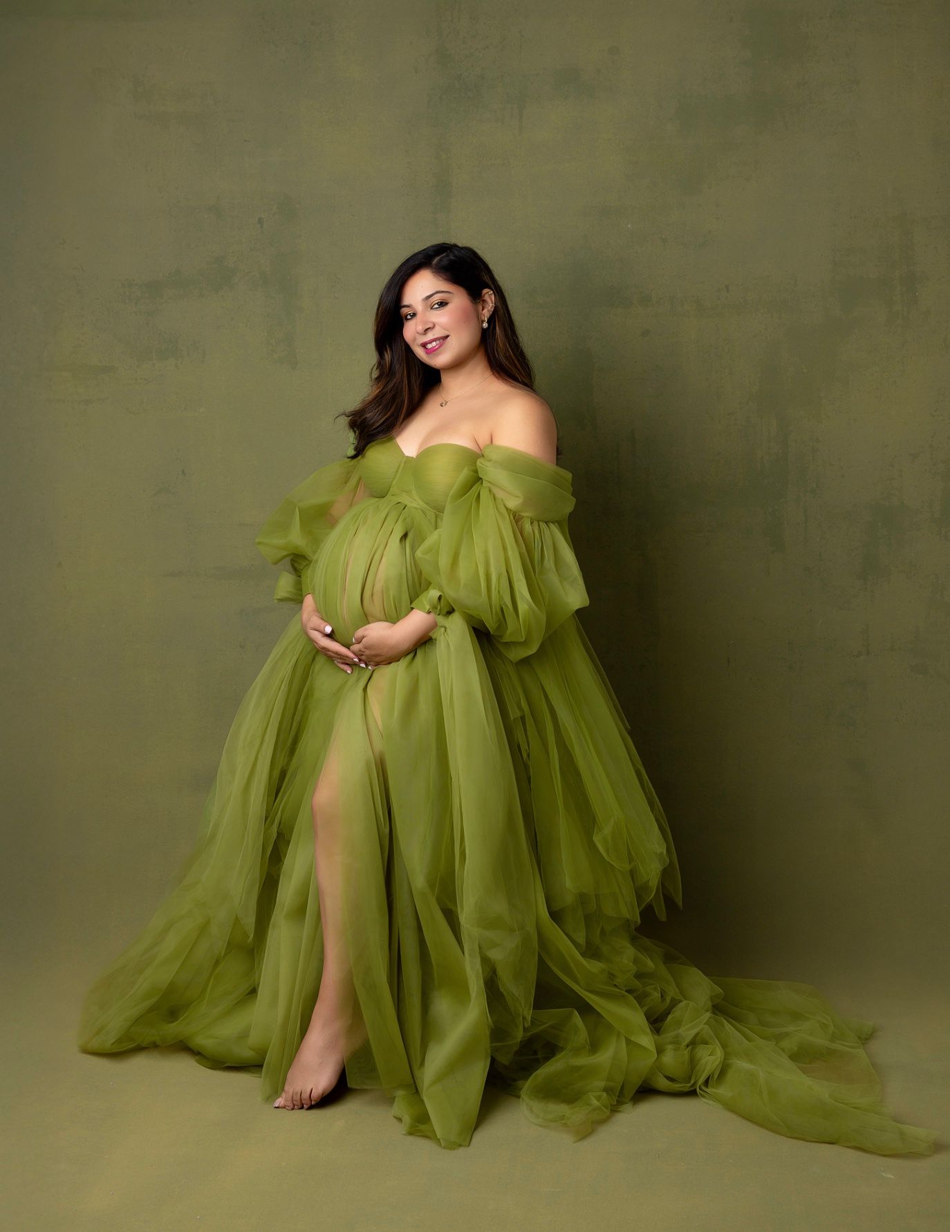 A flowing olive green tulle gown creates an ethereal silhouette against a textured sage backdrop.