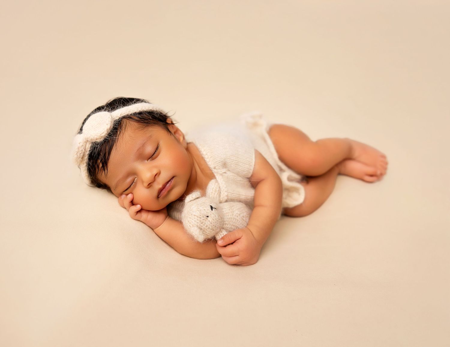 Sleeping newborn baby girl in white outfit holding a tiny stuffed bunny toy on a cream background.