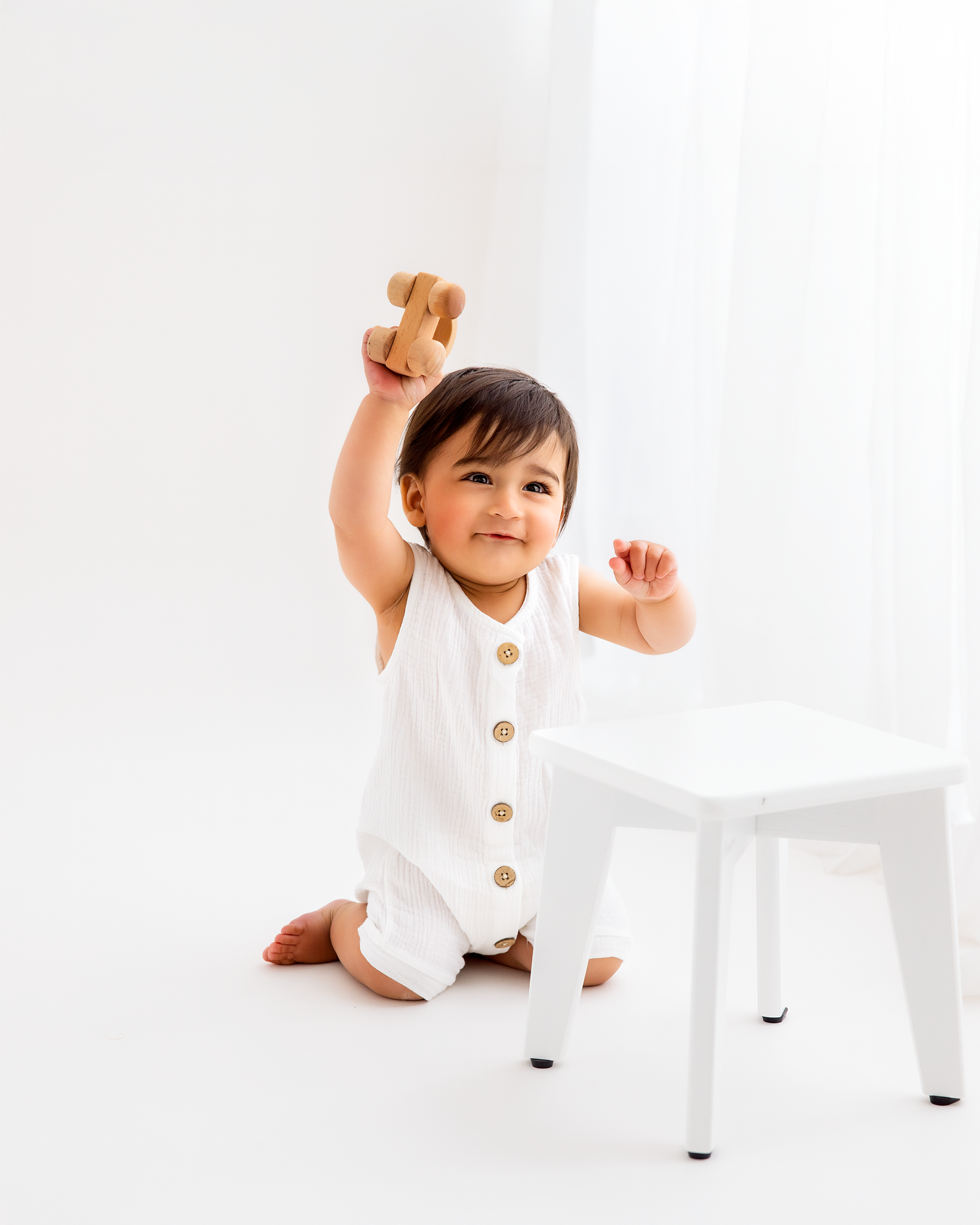 Smiling baby in white romper sits on floor, joyfully raising a wooden toy airplane above their head.