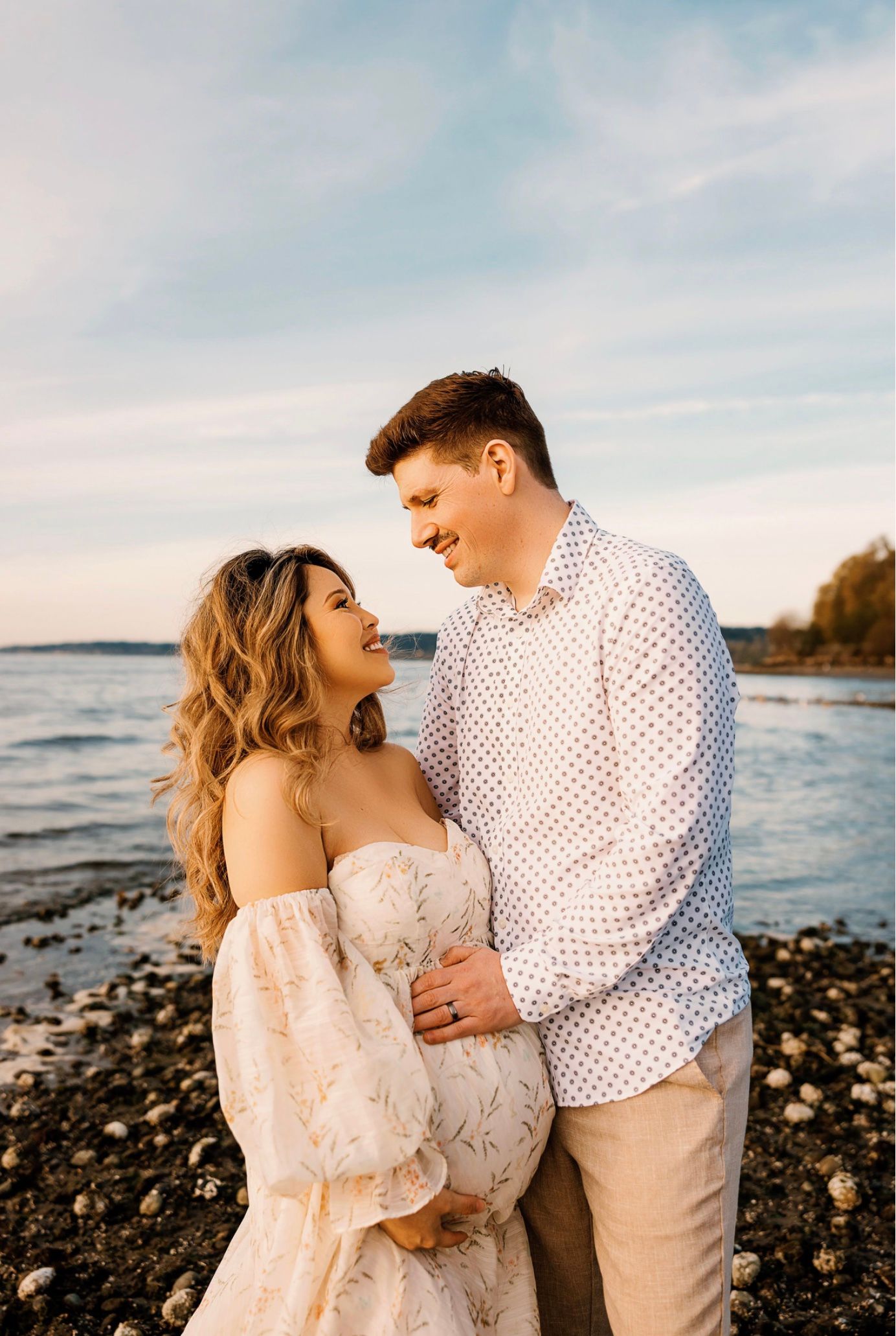 Couple sharing a tender moment on a rocky beach at golden hour, woman in floral maternity gown, man in polka dot shirt.