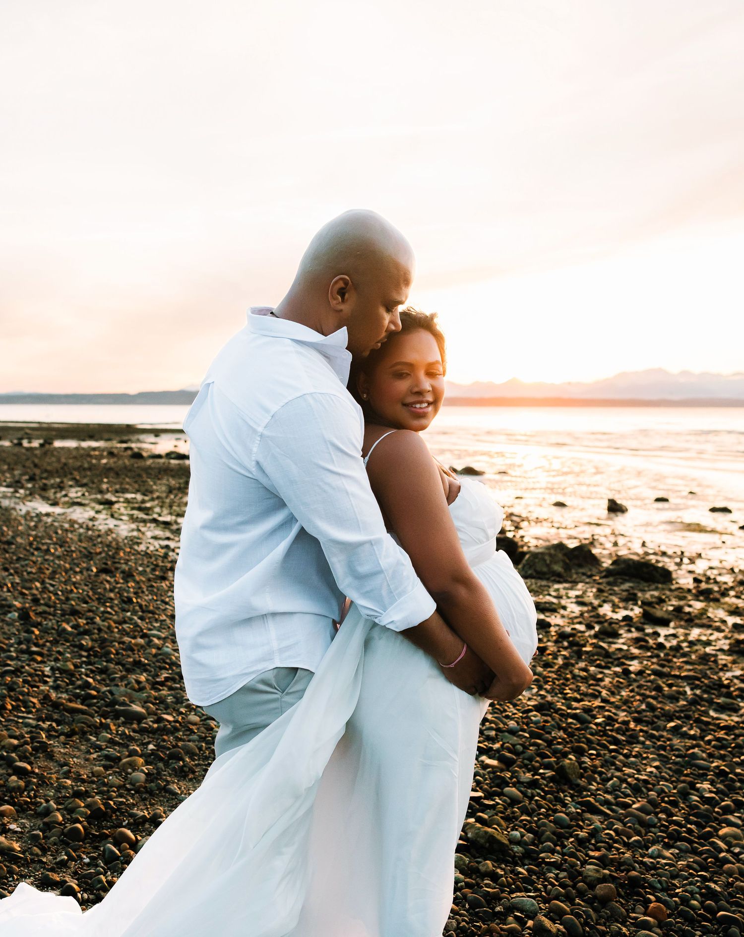 A couple embraces on a rocky beach at sunset, the woman in a white dress smiling as her partner holds her close.