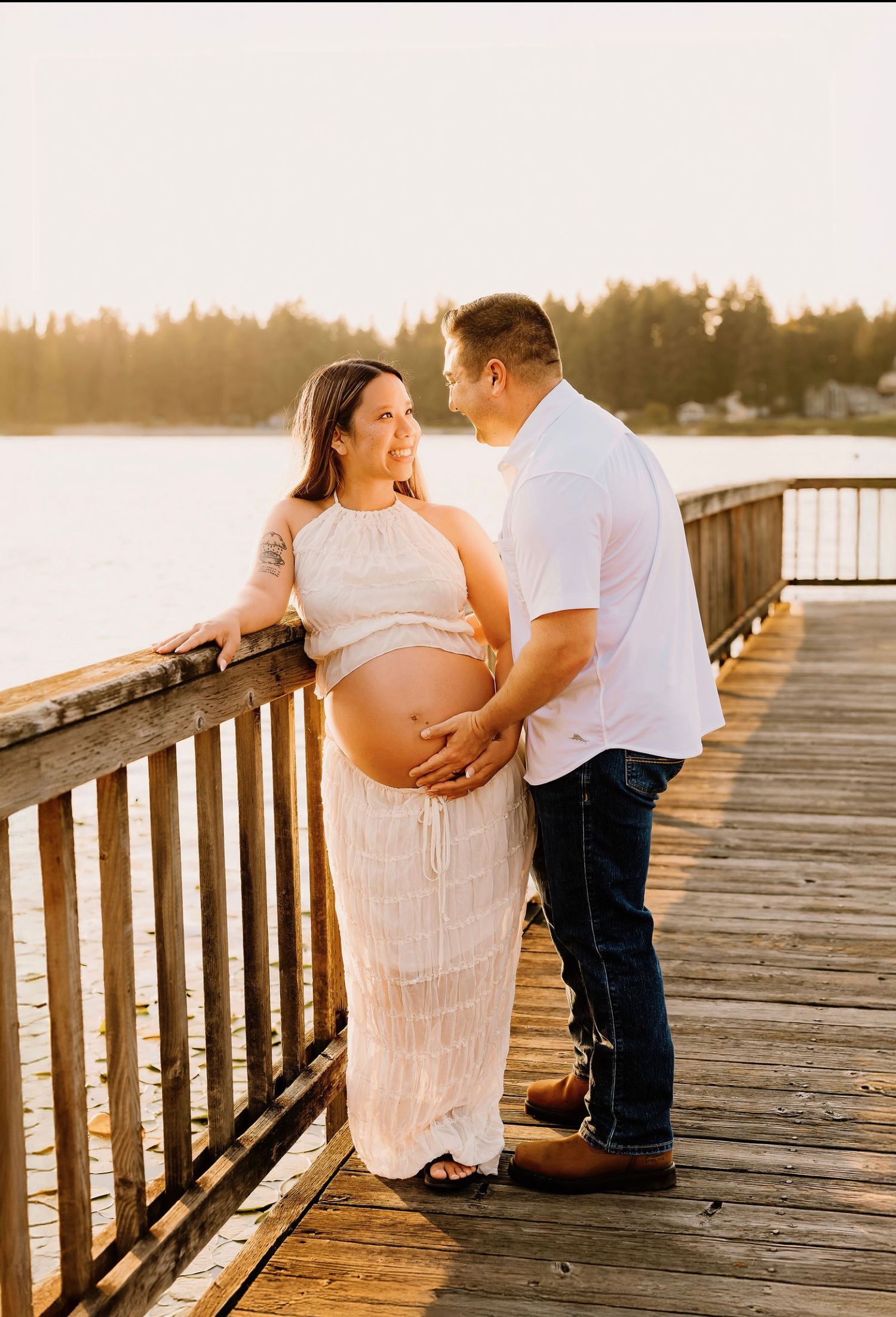 Pregnant couple sharing a tender moment on a wooden pier at golden hour, with a serene lake and forest backdrop.