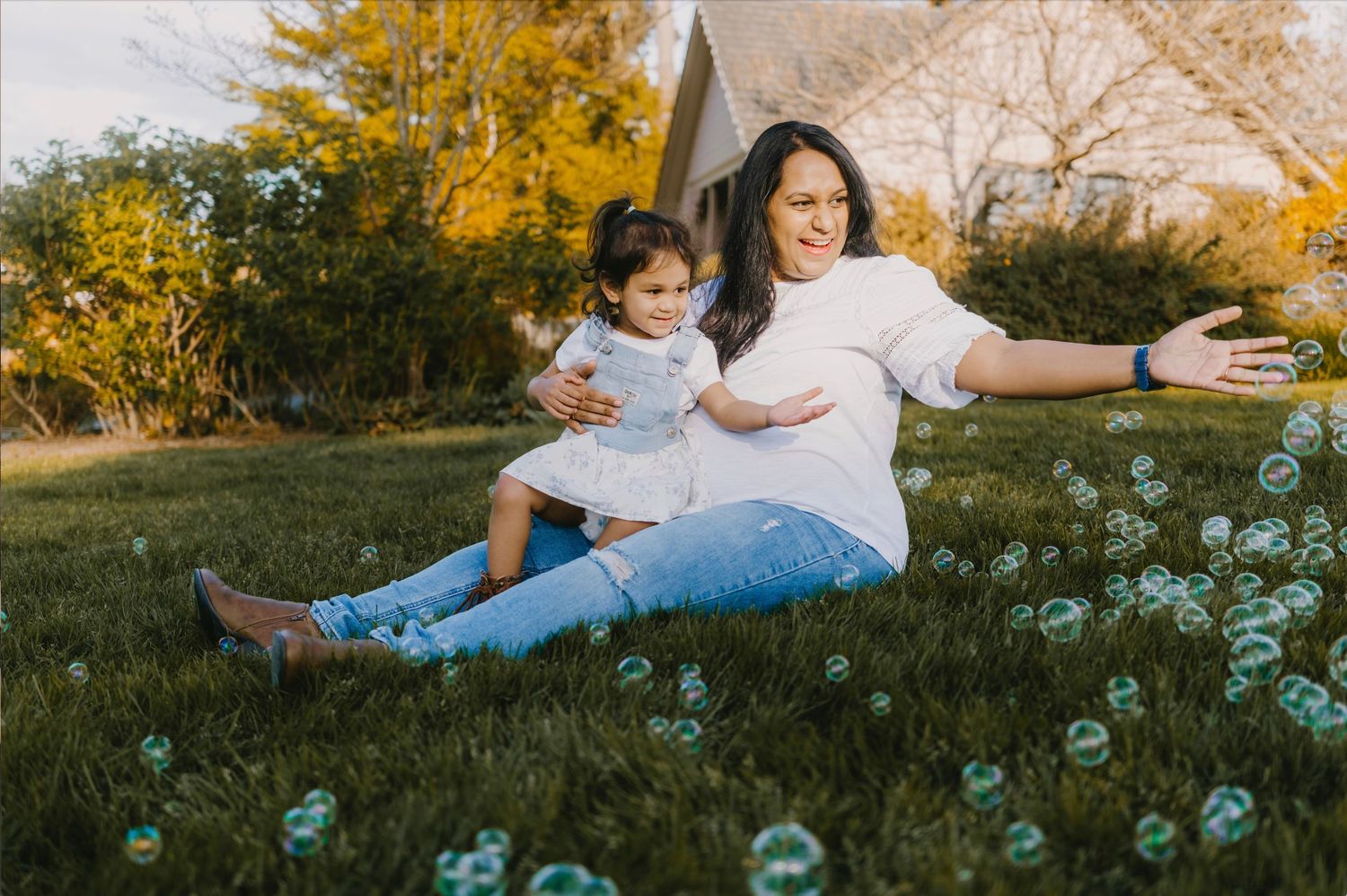 Pregnant woman sitting on grass with toddler girl, playing with bubbles outdoors in a sunny backyard.