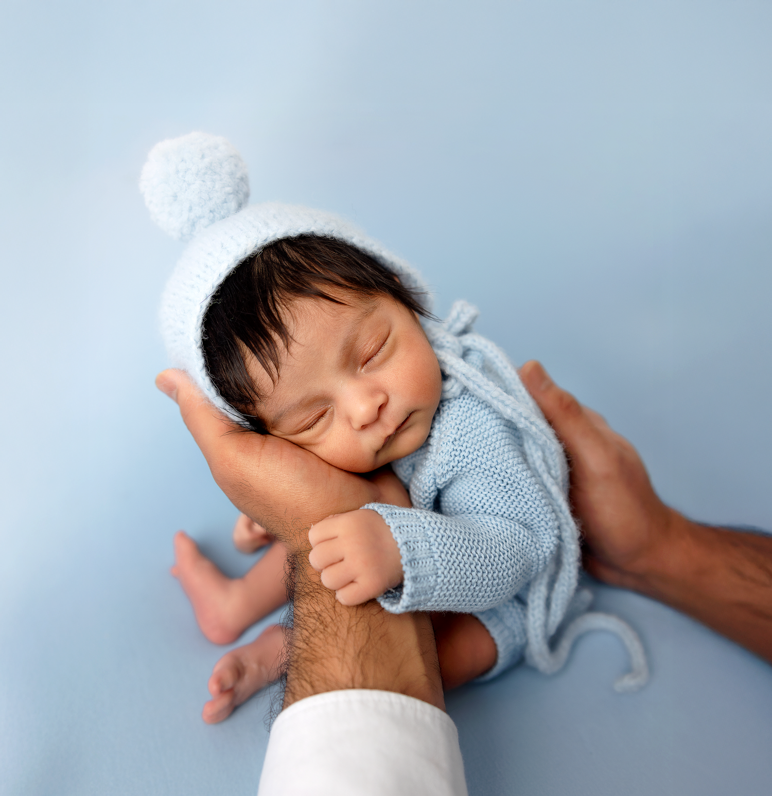Sleeping baby in blue knit sweater rests peacefully on white bedding with gentle hand caressing their head.