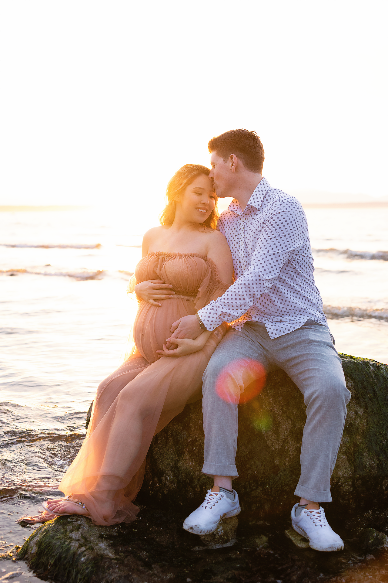 A couple sits together on coastal rocks during sunset creating a romantic silhouette against the golden light.