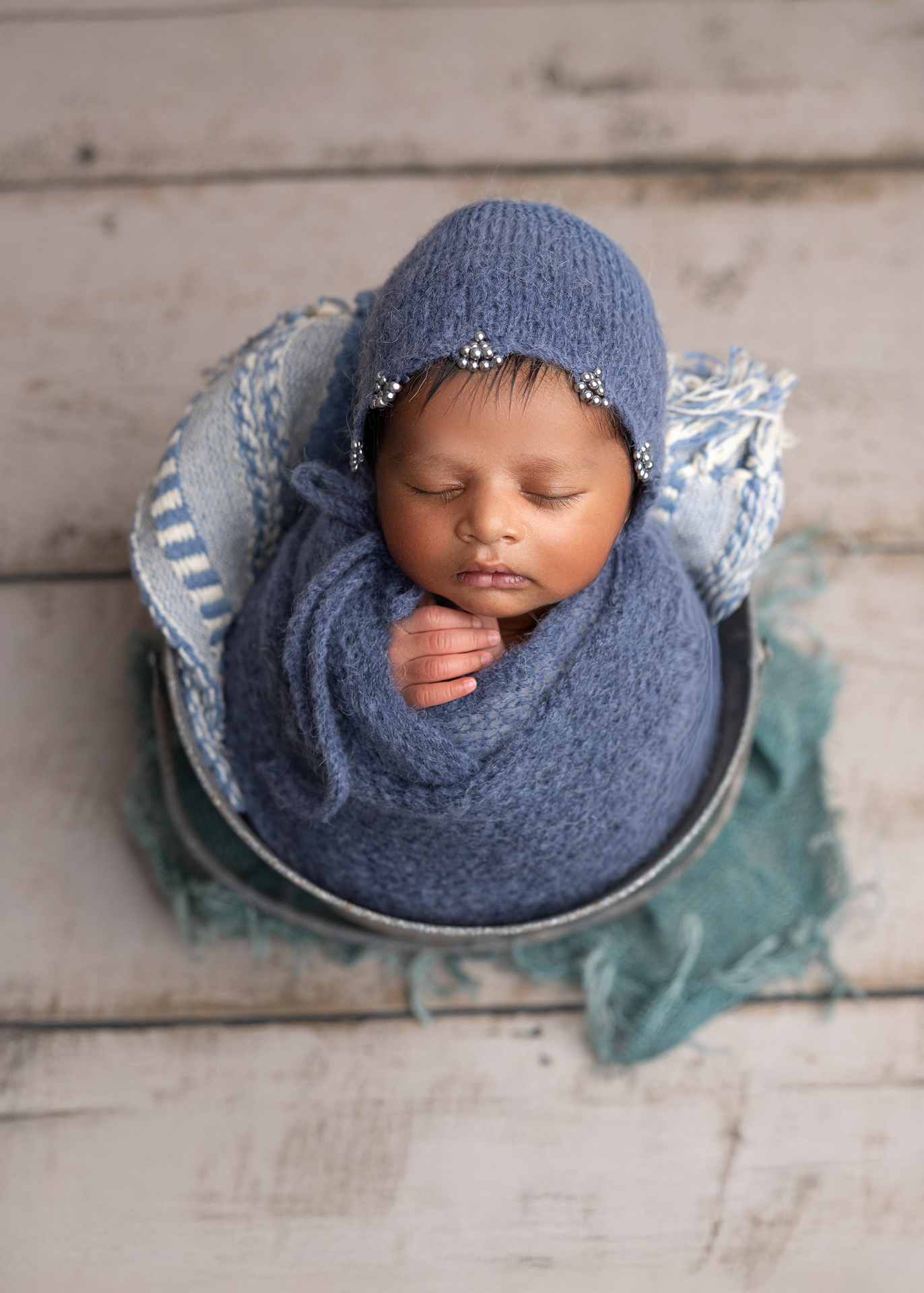 A sleeping baby swaddled in navy blue knit fabric lies in a teal-colored bowl against a rustic wooden backdrop.