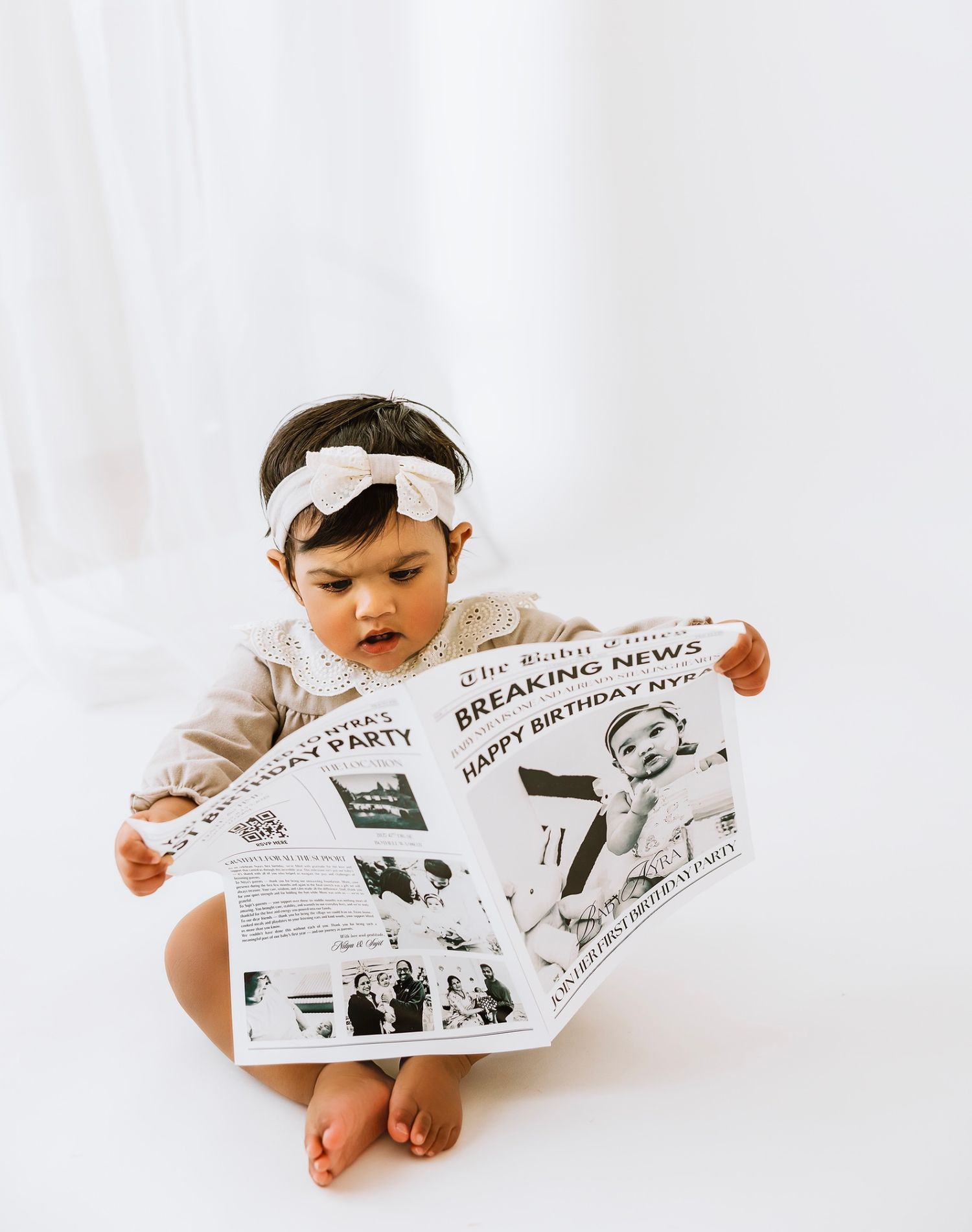 A person in white clothing sits cross-legged while reading a newspaper spread out in front of them.