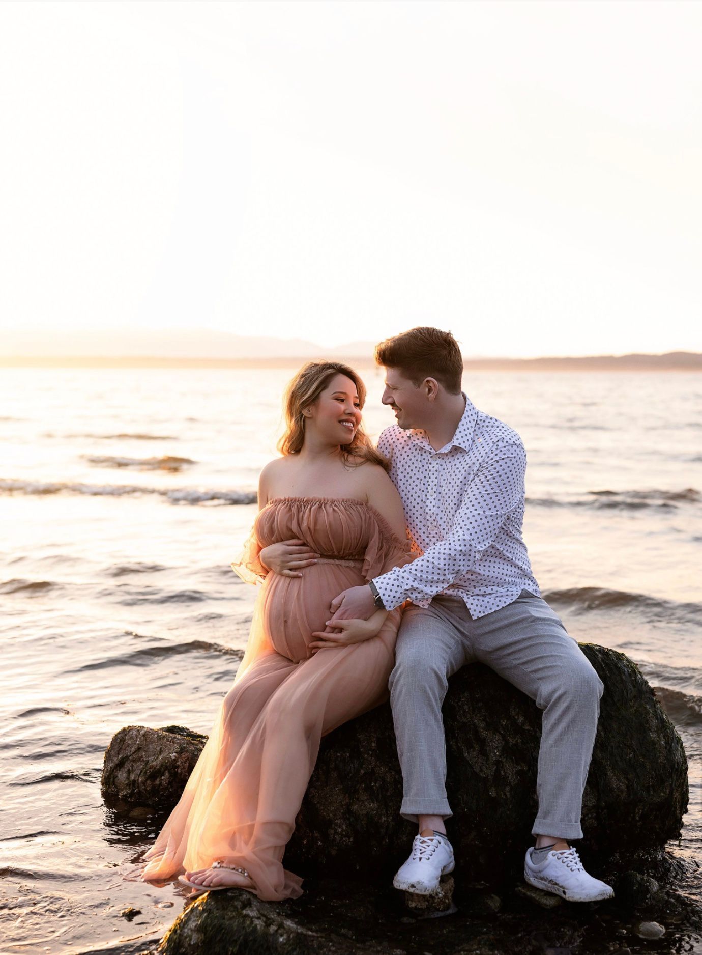 Couple sharing a tender moment on a rock by the ocean at sunset, woman in pink maternity gown cradling her bump.
