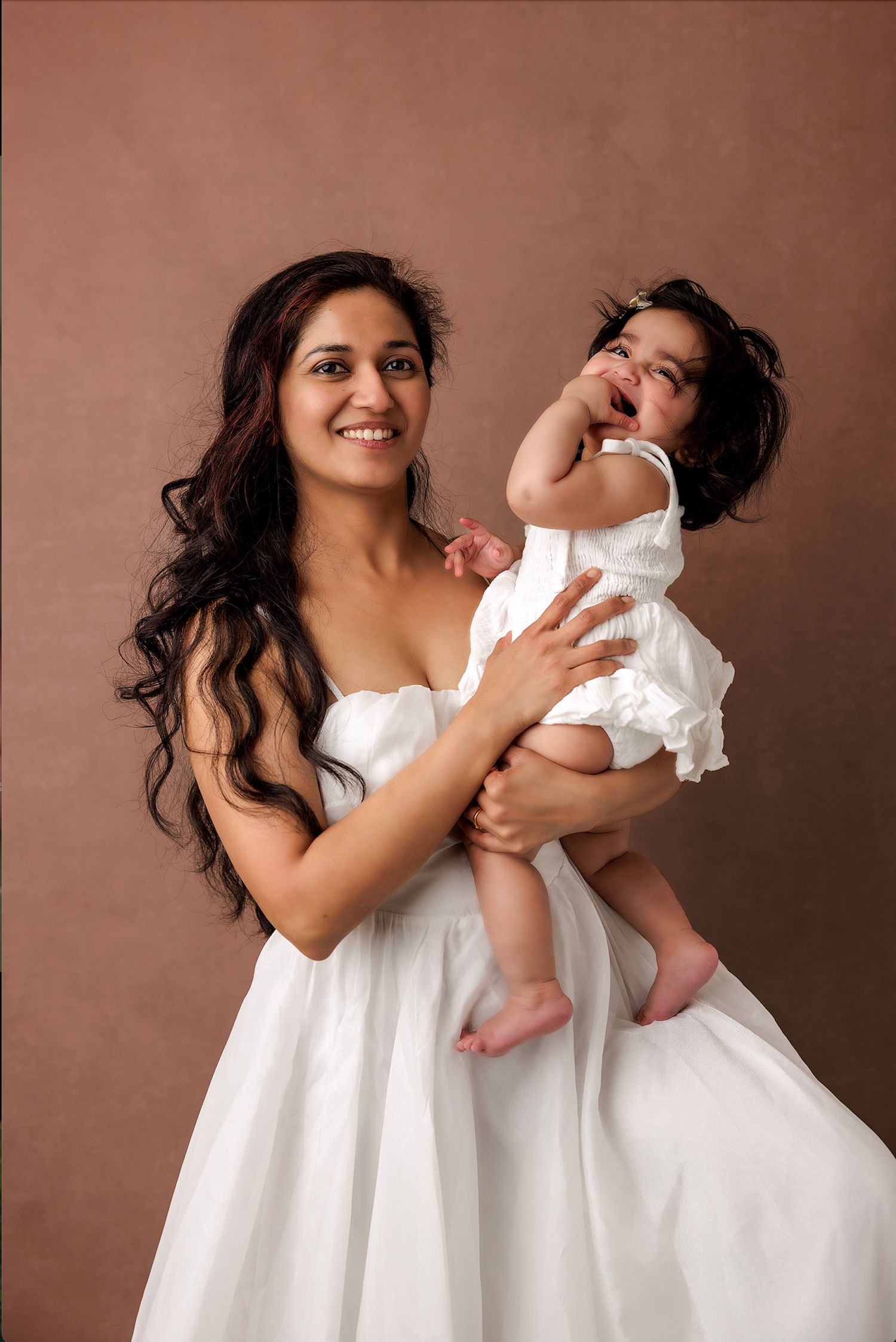 Joyful portrait of parent holding laughing baby in white dress.