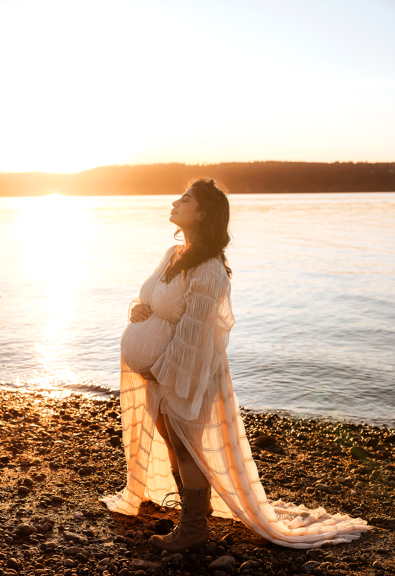Pregnant woman in flowing white dress stands on rocky beach at golden sunset by calm water.