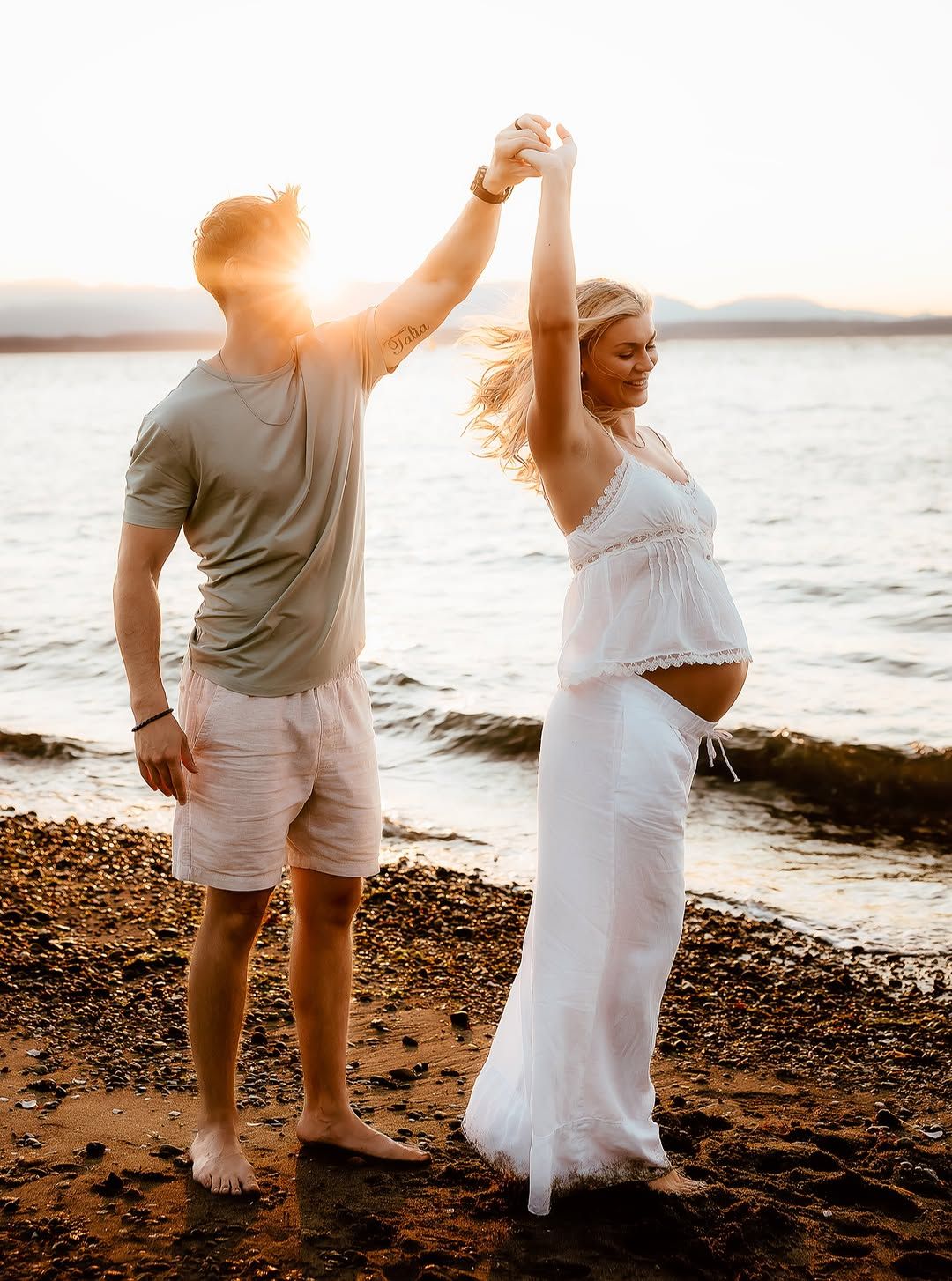 Pregnant couple dancing barefoot on a rocky beach at golden sunset, woman in white maternity dress.