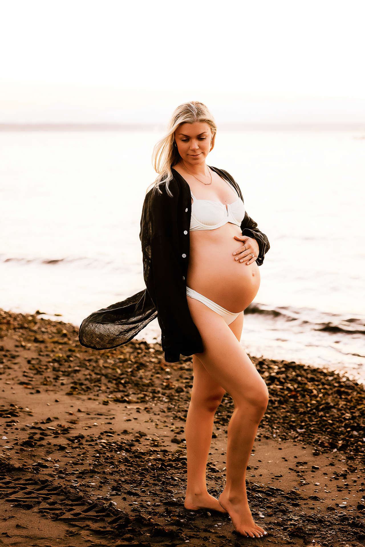 Pregnant woman in white bikini and black sheer shirt cradling baby bump on rocky beach at sunset.
