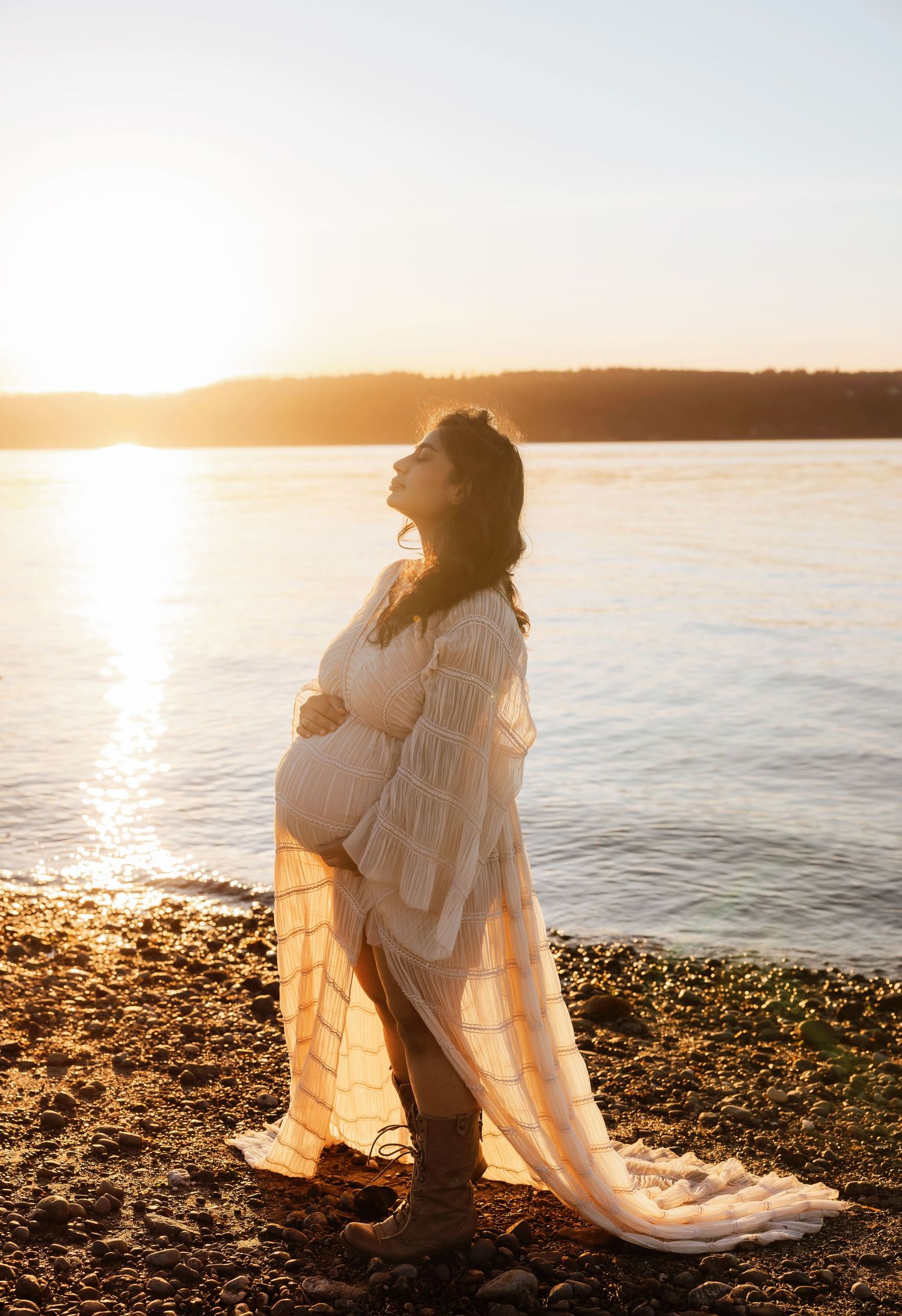 A dreamy coastal photoshoot at golden hour shows a figure in white sitting by the rocky shore as sunlight sparkles on the water.