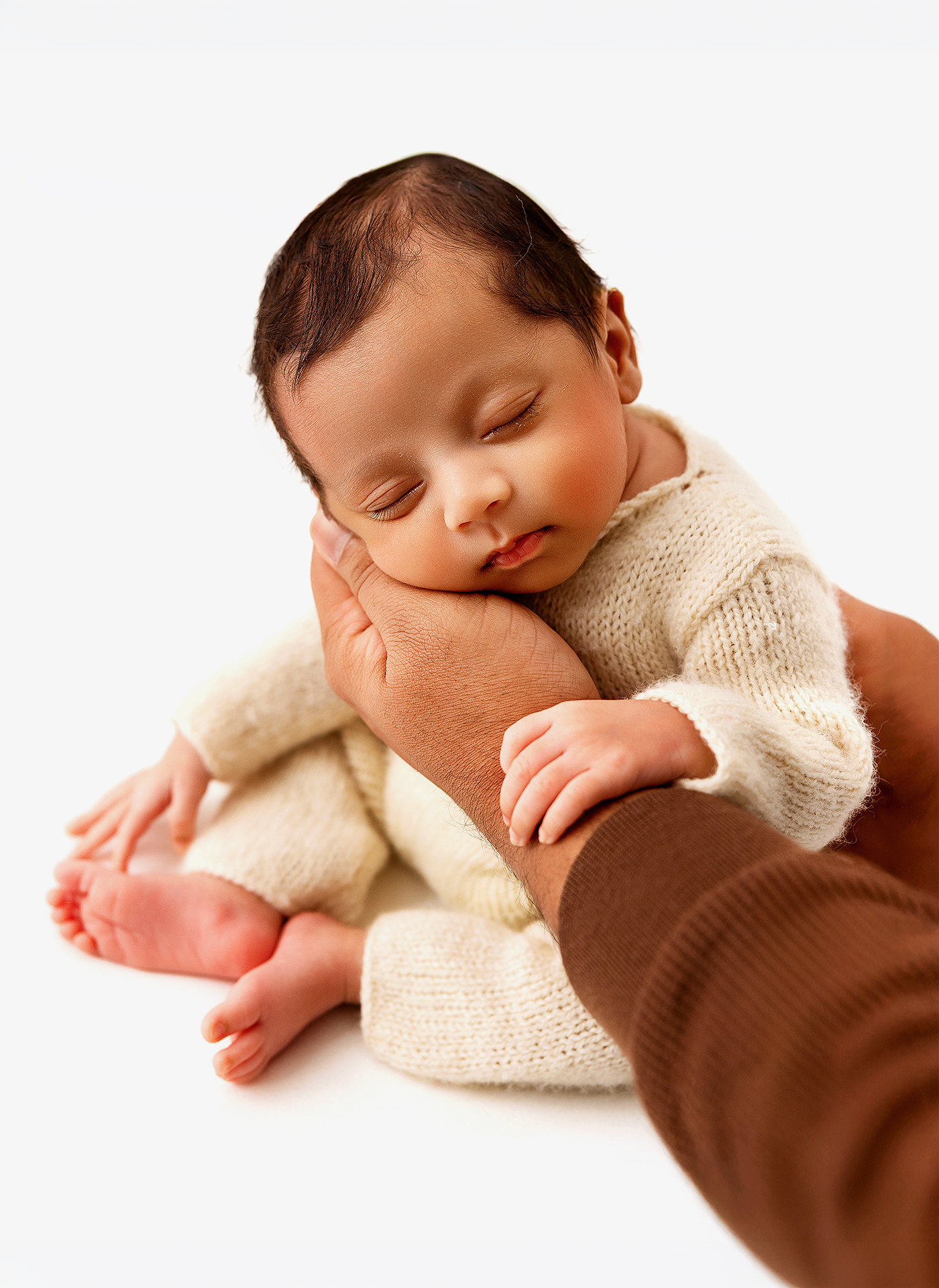 A sleeping newborn baby in a cream knit outfit rests peacefully in an adult's supportive hands against white background.