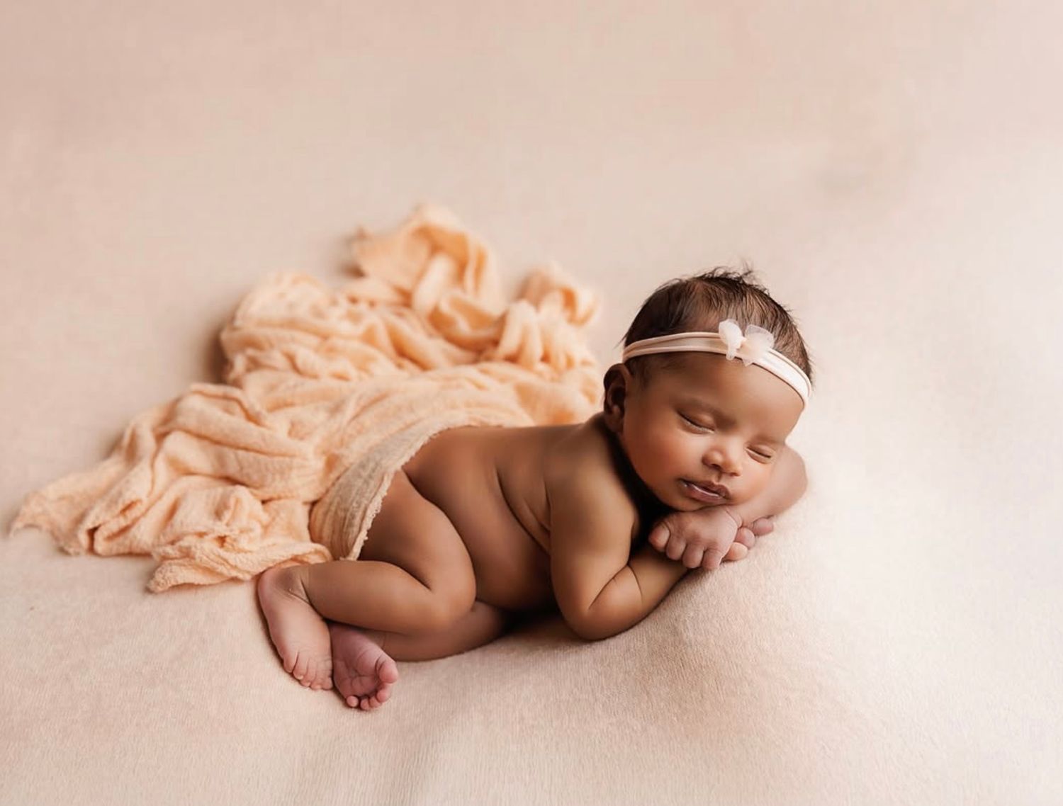 Peaceful newborn baby sleeping on a soft peach blanket, wearing a delicate floral headband in a professional photo.