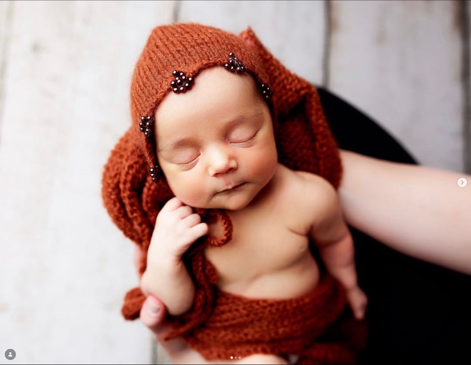 Sleeping newborn baby wearing a rust orange knit bonnet with black bead accents, cradled in a photographer's hands.