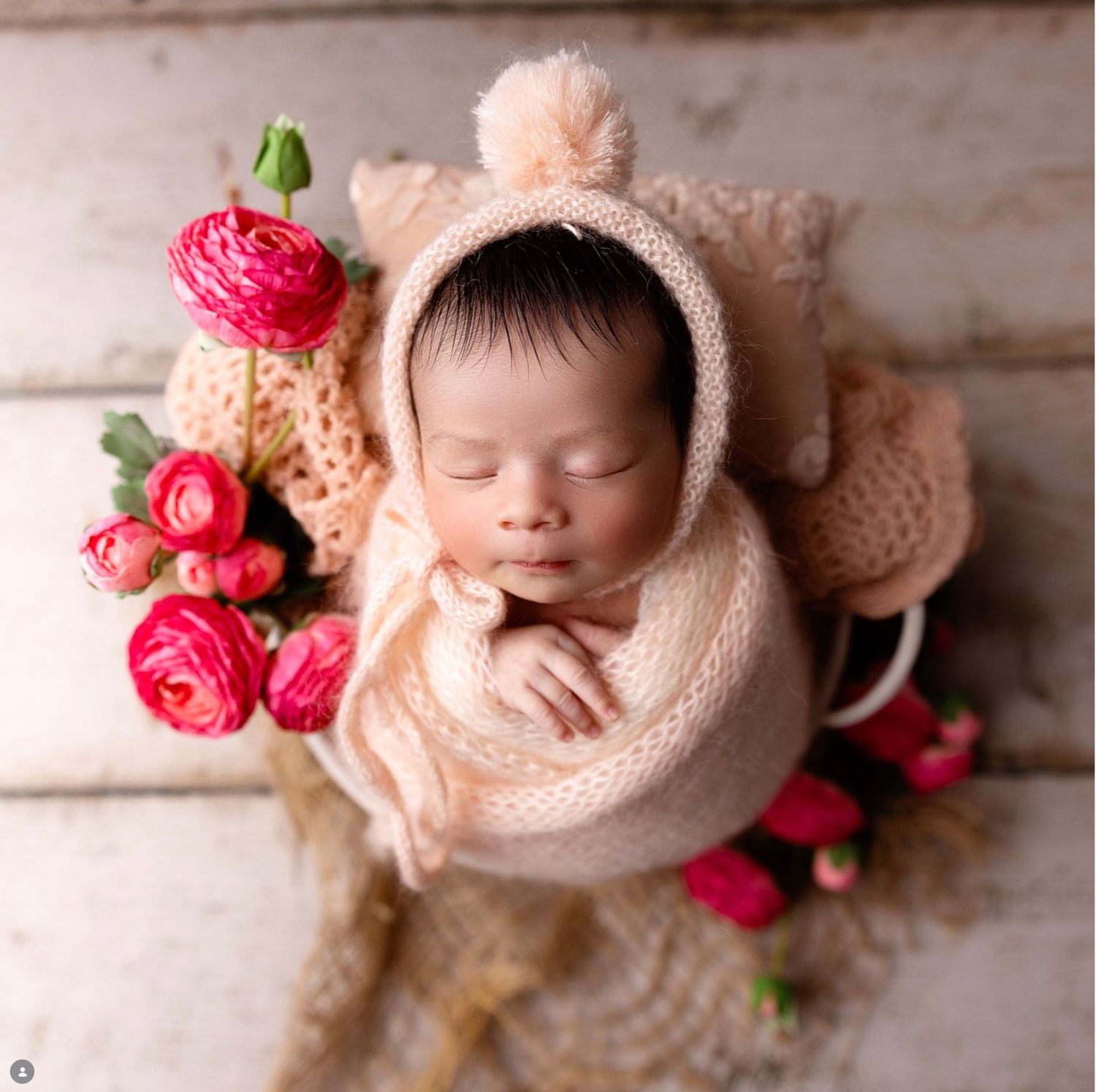 A sleeping newborn swaddled in soft peach fabric with pink ranunculus flowers in a dreamy studio portrait.