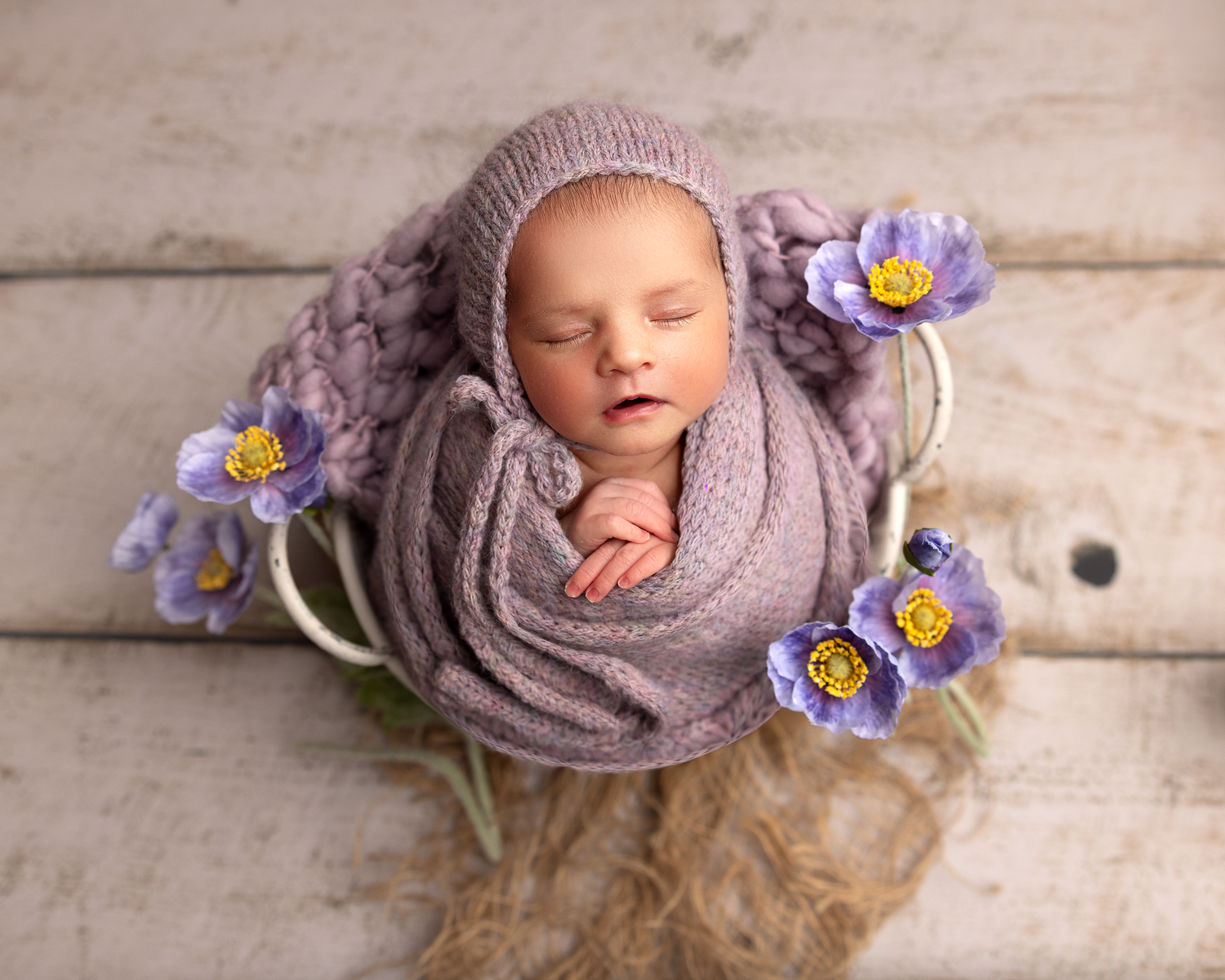 Sleeping baby wrapped in soft purple knit blanket surrounded by delicate purple flowers on wooden backdrop.