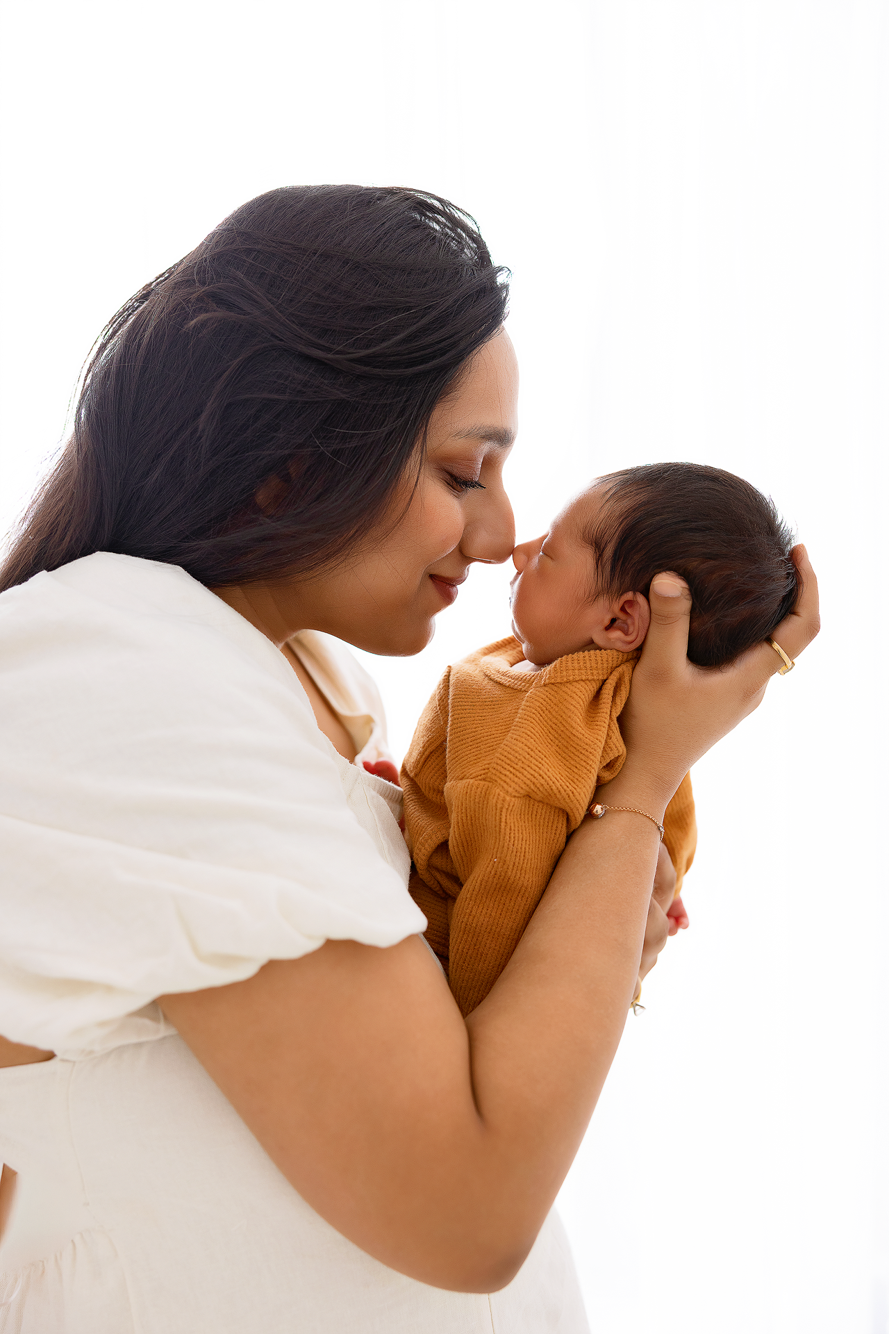 A tender moment between parent and infant as they touch noses, with the baby wearing a brown onesie against white background, Everett .