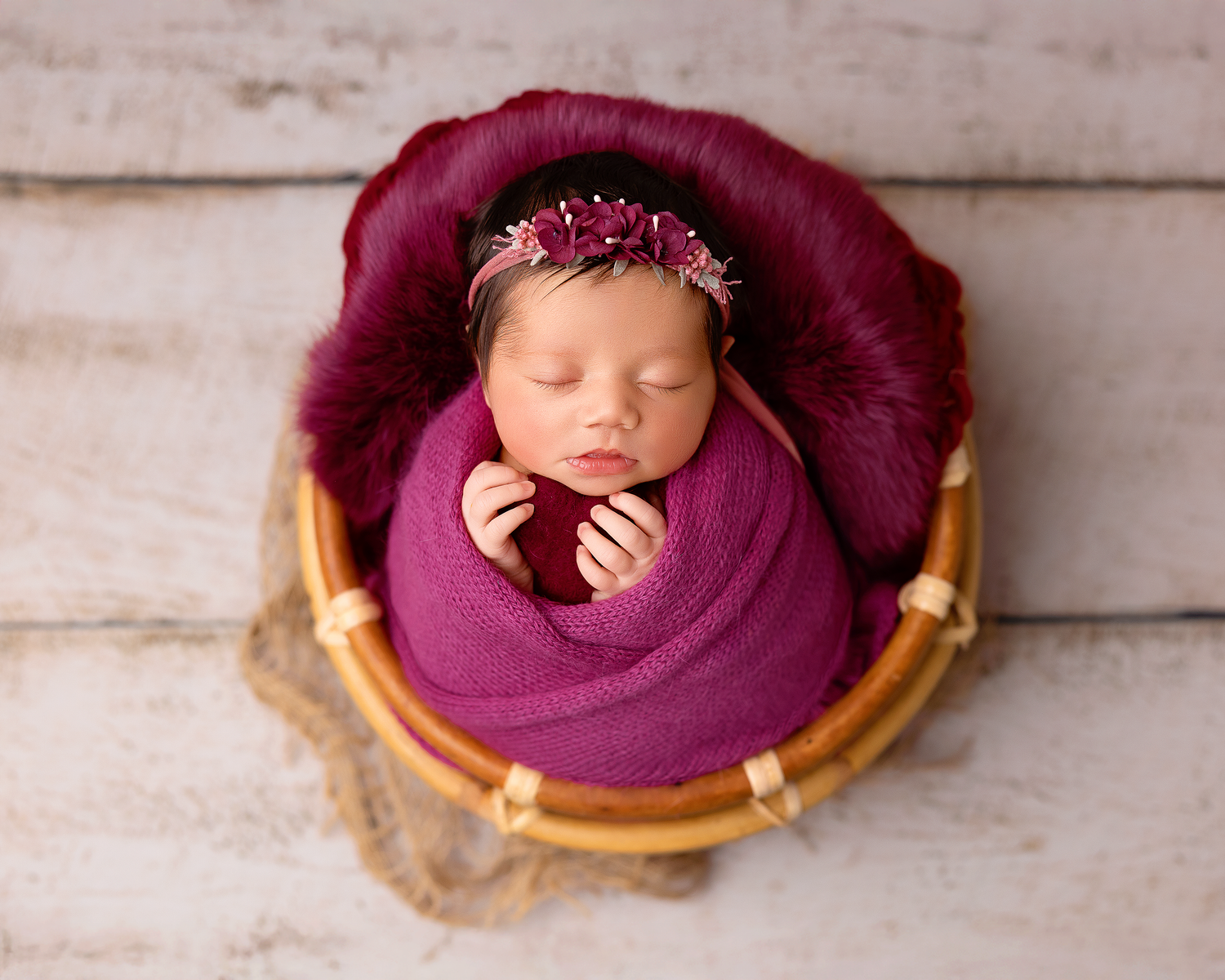 A newborn baby wrapped in berry fabric lies sleeping in a wooden bowl against a rustic white background.