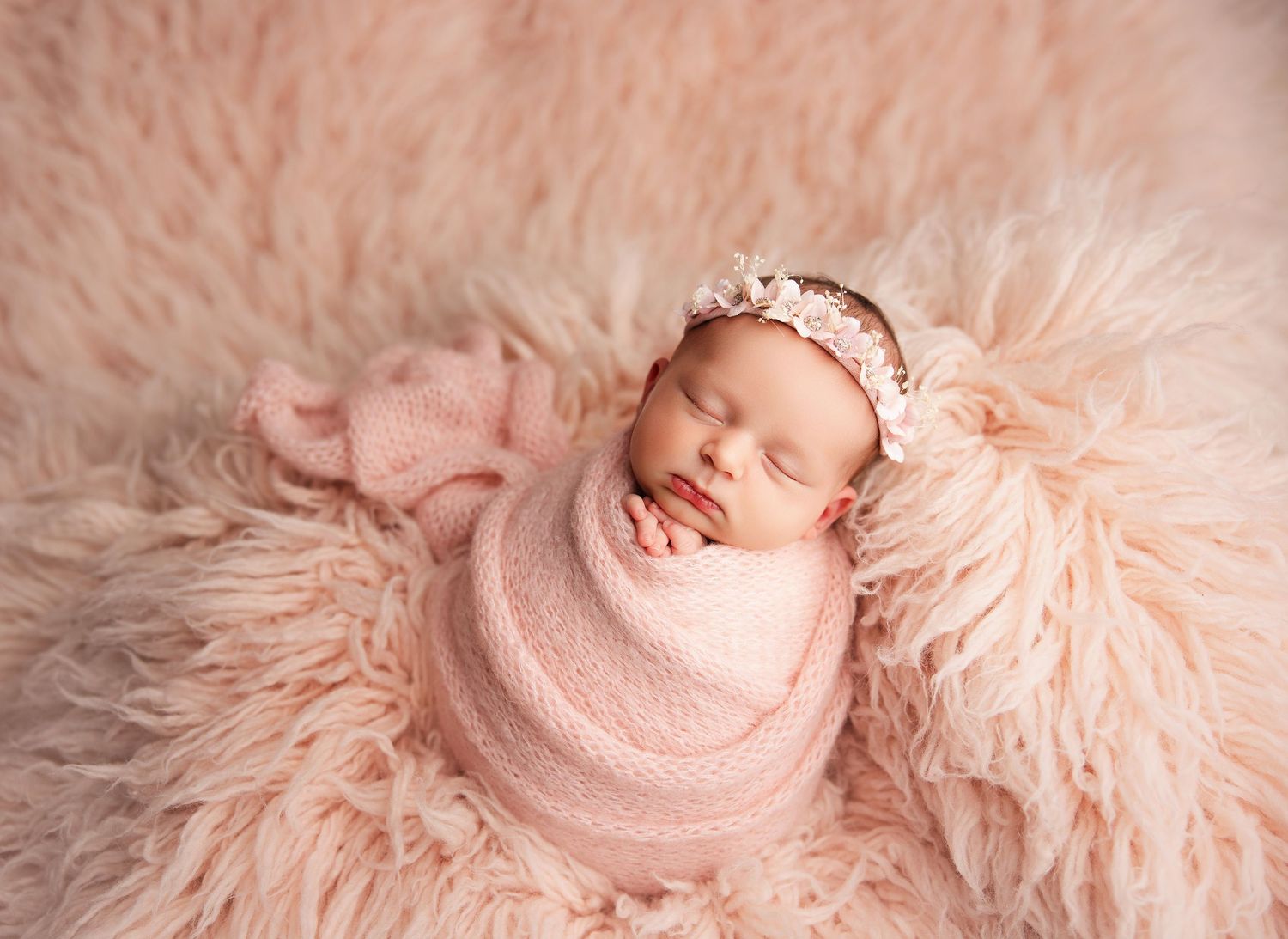 Sleeping newborn baby girl wrapped in pink swaddle with floral headband on fluffy pink fur blanket.
