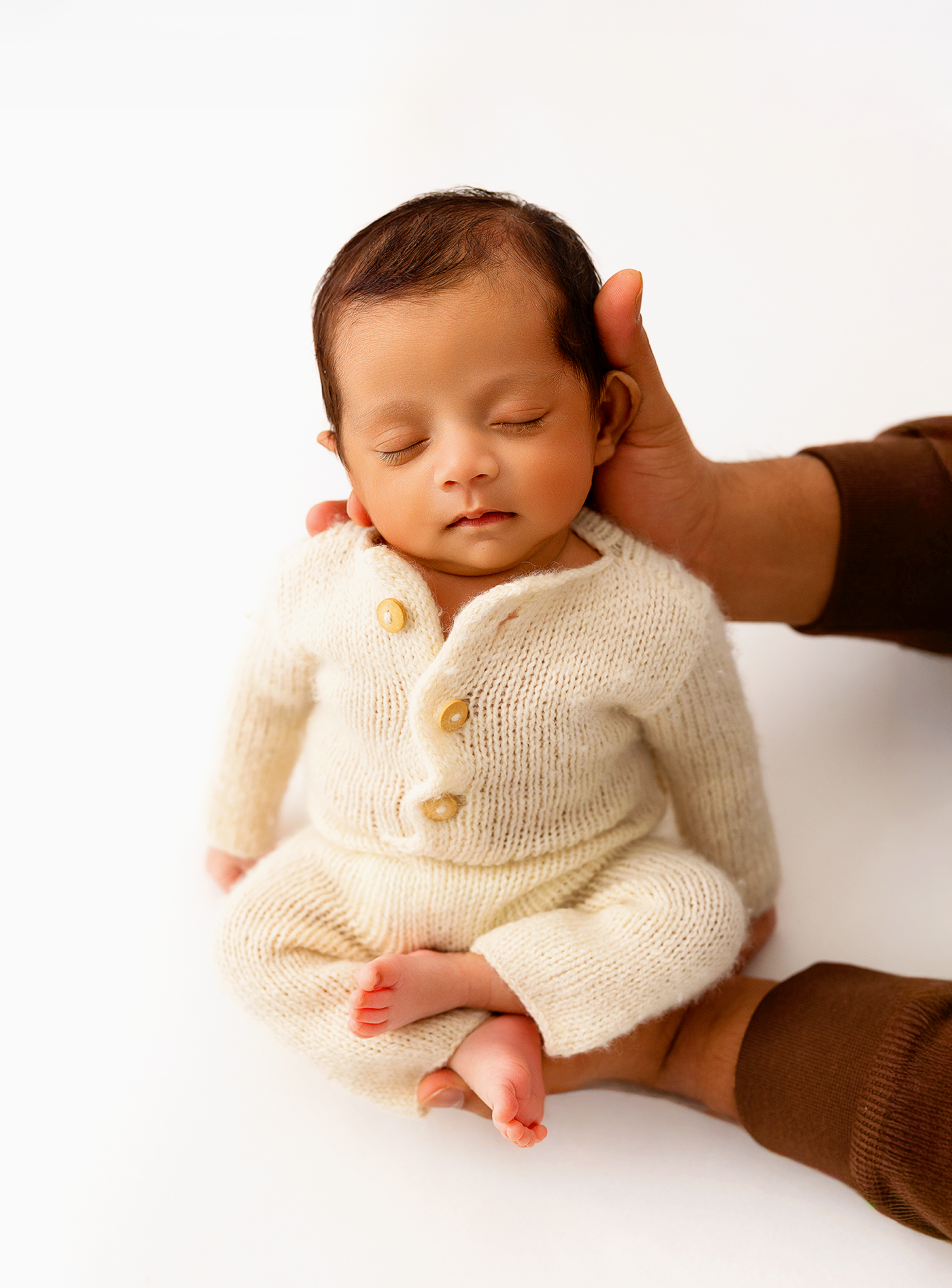 Sleeping newborn baby in cream knit romper cradled by parent hands on white background.