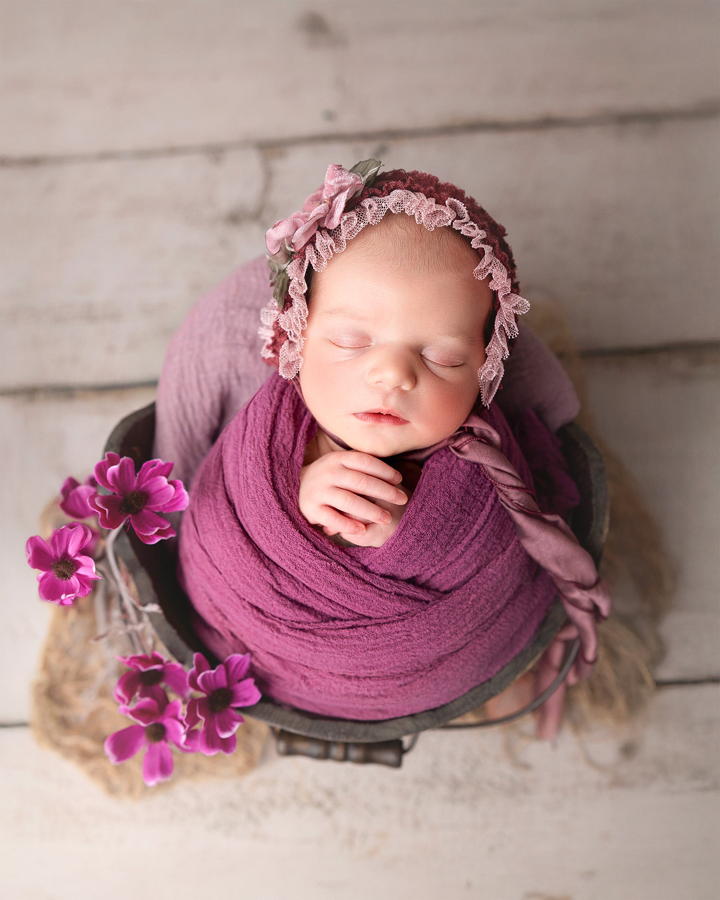 Newborn wrapped in purple fabric with matching bonnet and pink flowers resting peacefully on wooden floor.