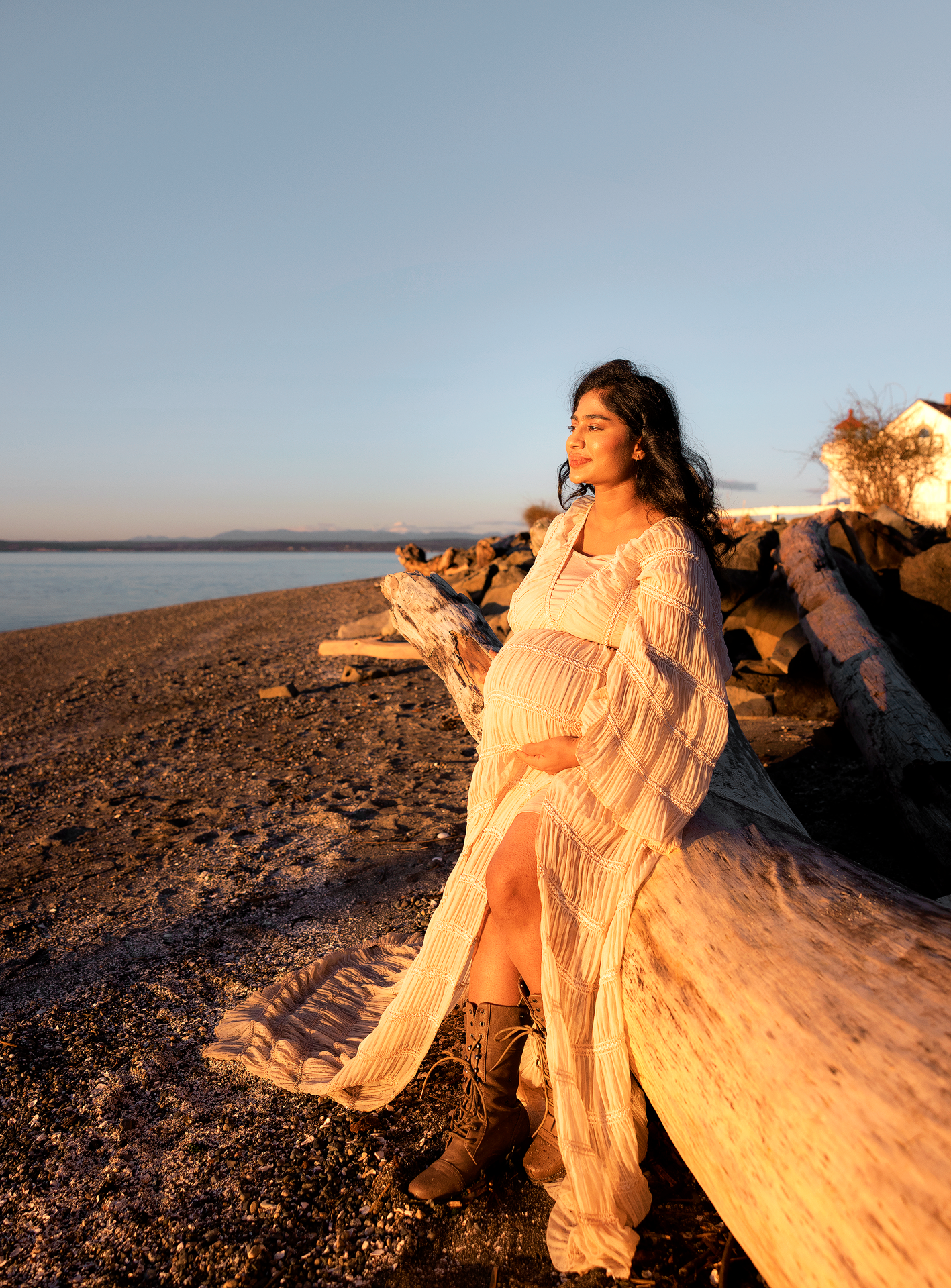 Pregnant woman in white lace dress and boots sits on driftwood at golden hour beach.