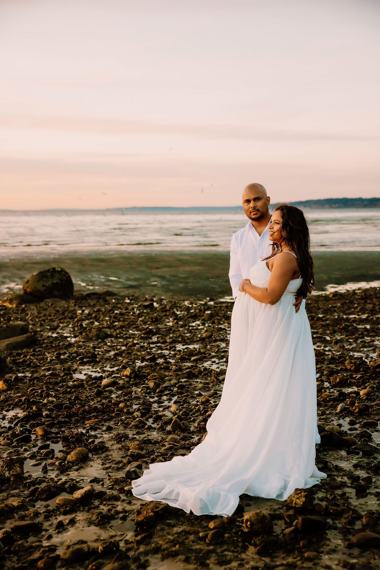 Couple in white attire embracing on rocky beach at sunset with calm ocean waves in the background.