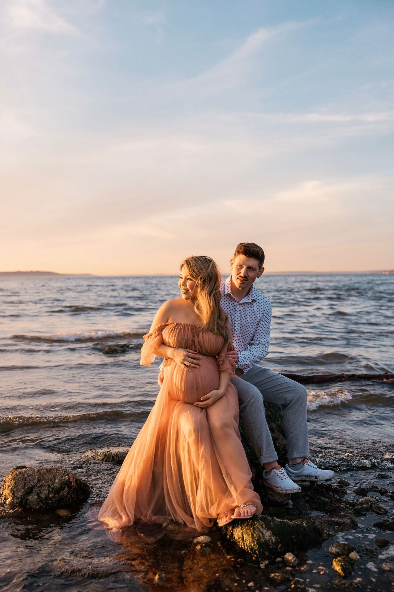 Pregnant couple poses on rocky shoreline at sunset, woman in flowing peach gown, man in casual shirt, ocean backdrop.