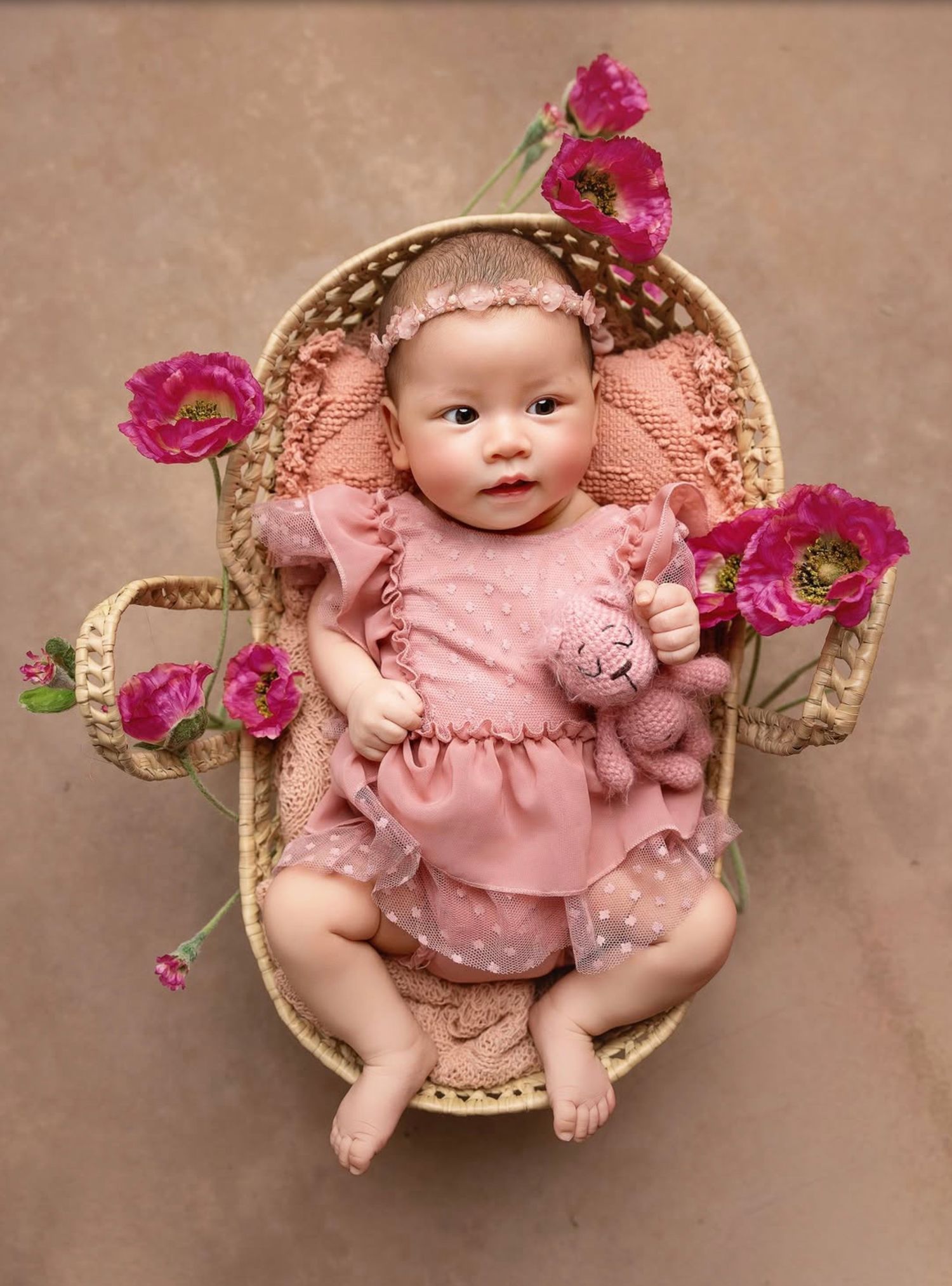 A baby in a pink lace dress lies in a woven basket decorated with vibrant pink flowers against a brown backdrop.