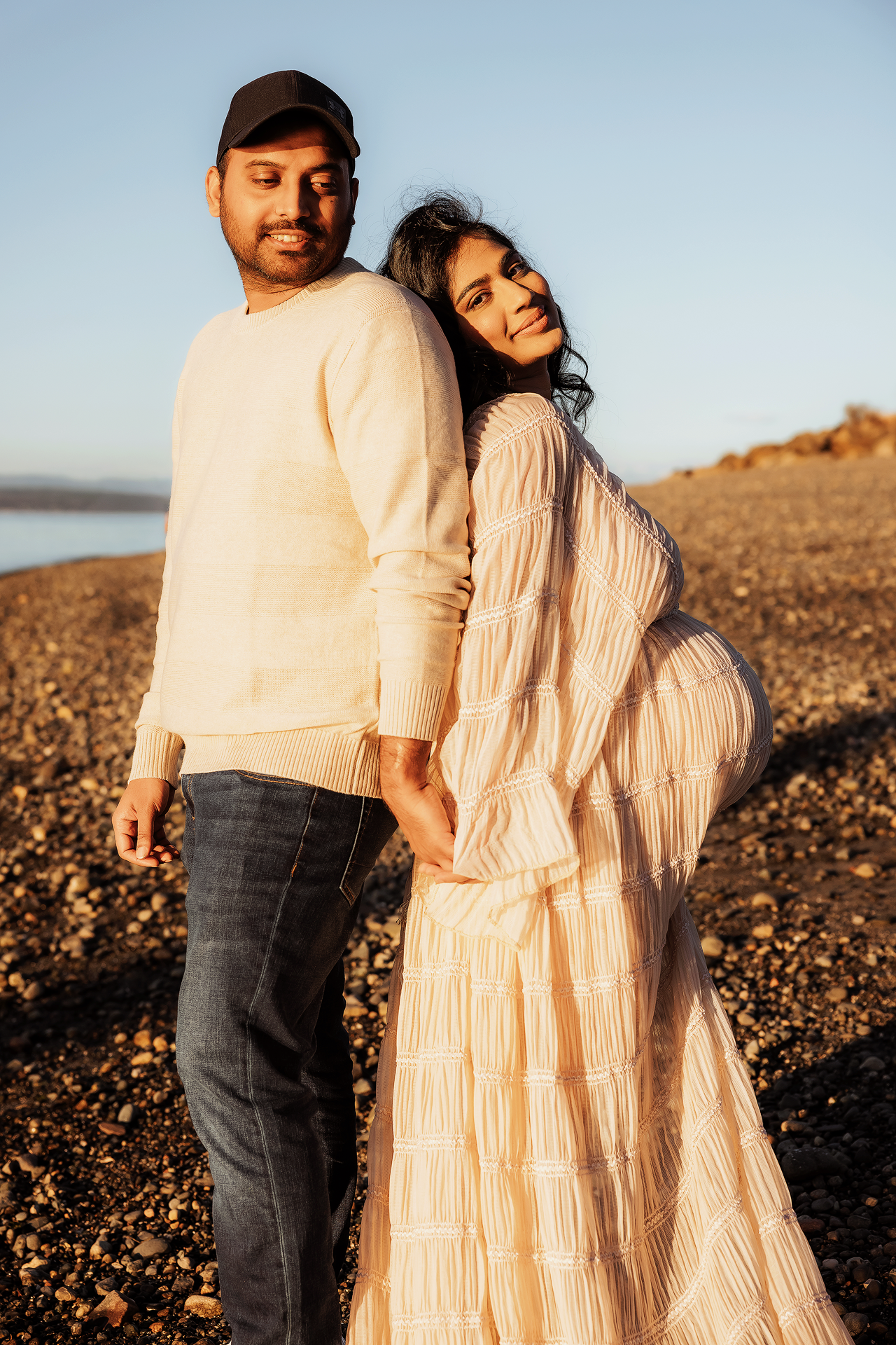 A couple stands together on rocky terrain at sunset wearing light colored clothing and embracing each other.