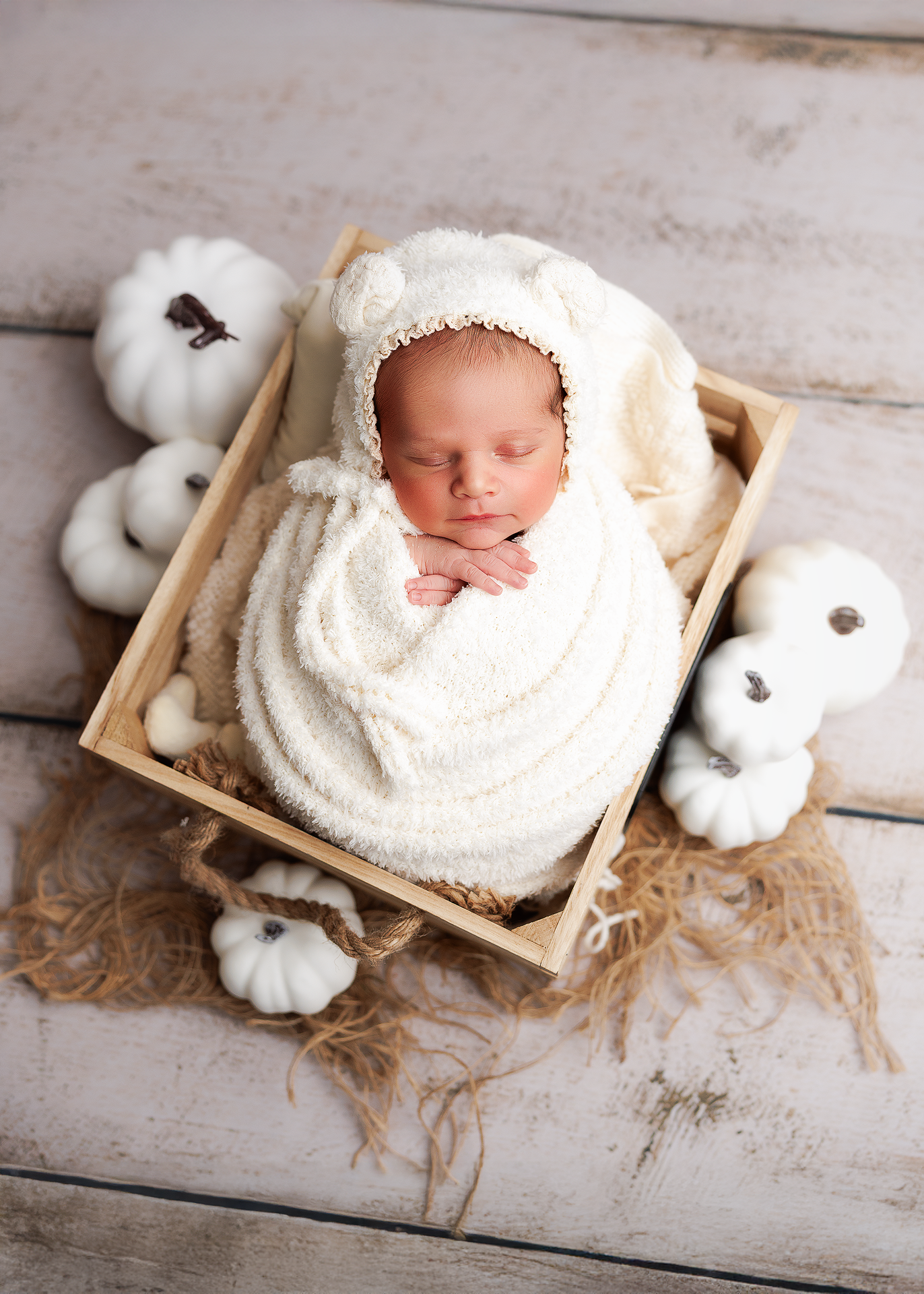 A sleeping newborn swaddled in white blankets rests in a rustic wooden crate surrounded by cotton pumpkins on wooden floor.