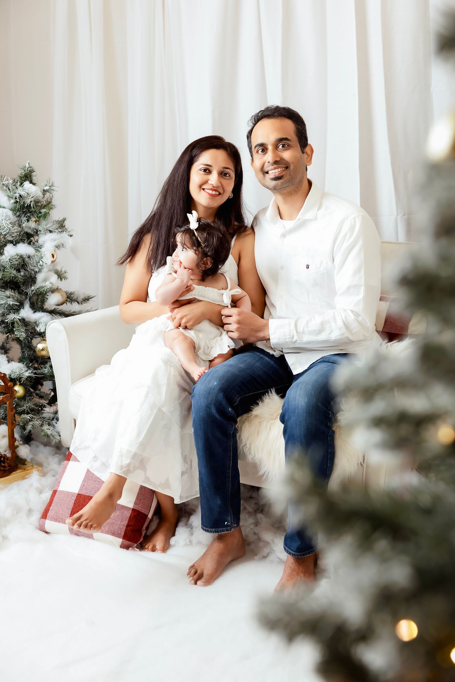 Happy family with baby girl posing by Christmas trees in white studio setting for holiday portrait.