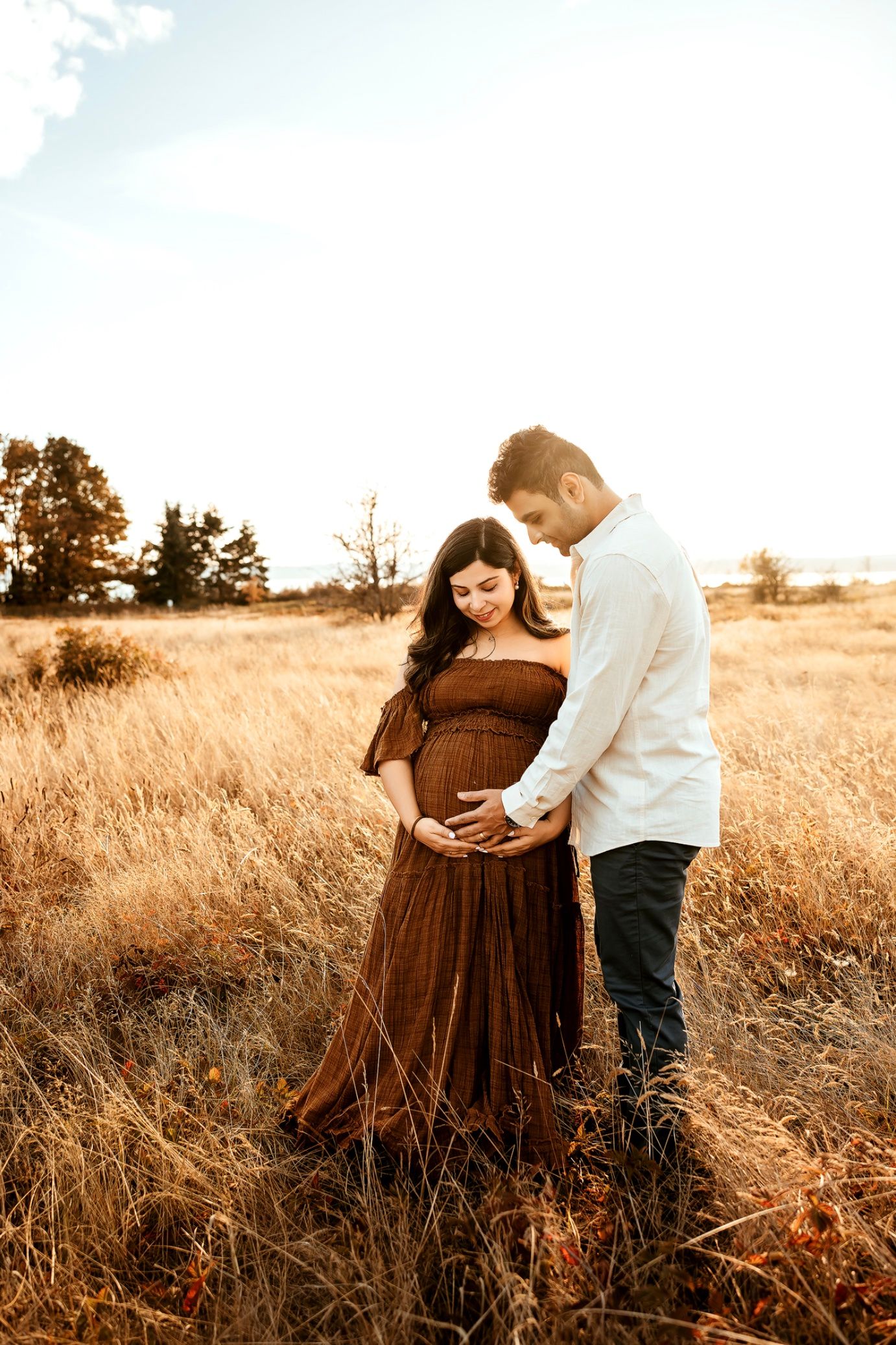 Couple in golden field, pregnant woman in brown dress, partner in white shirt, both looking at baby bump at sunset.
