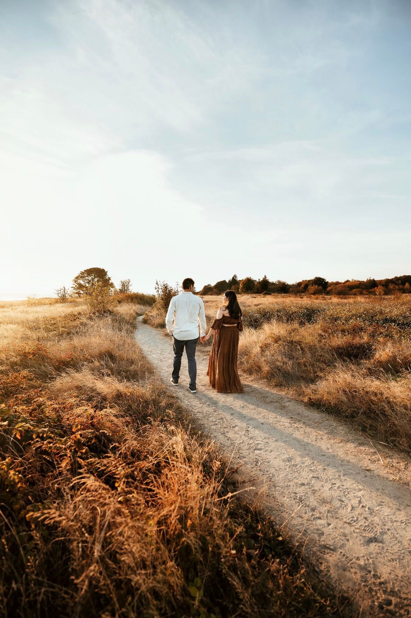 Couple holding hands walking down a dirt path through golden fields at sunset.