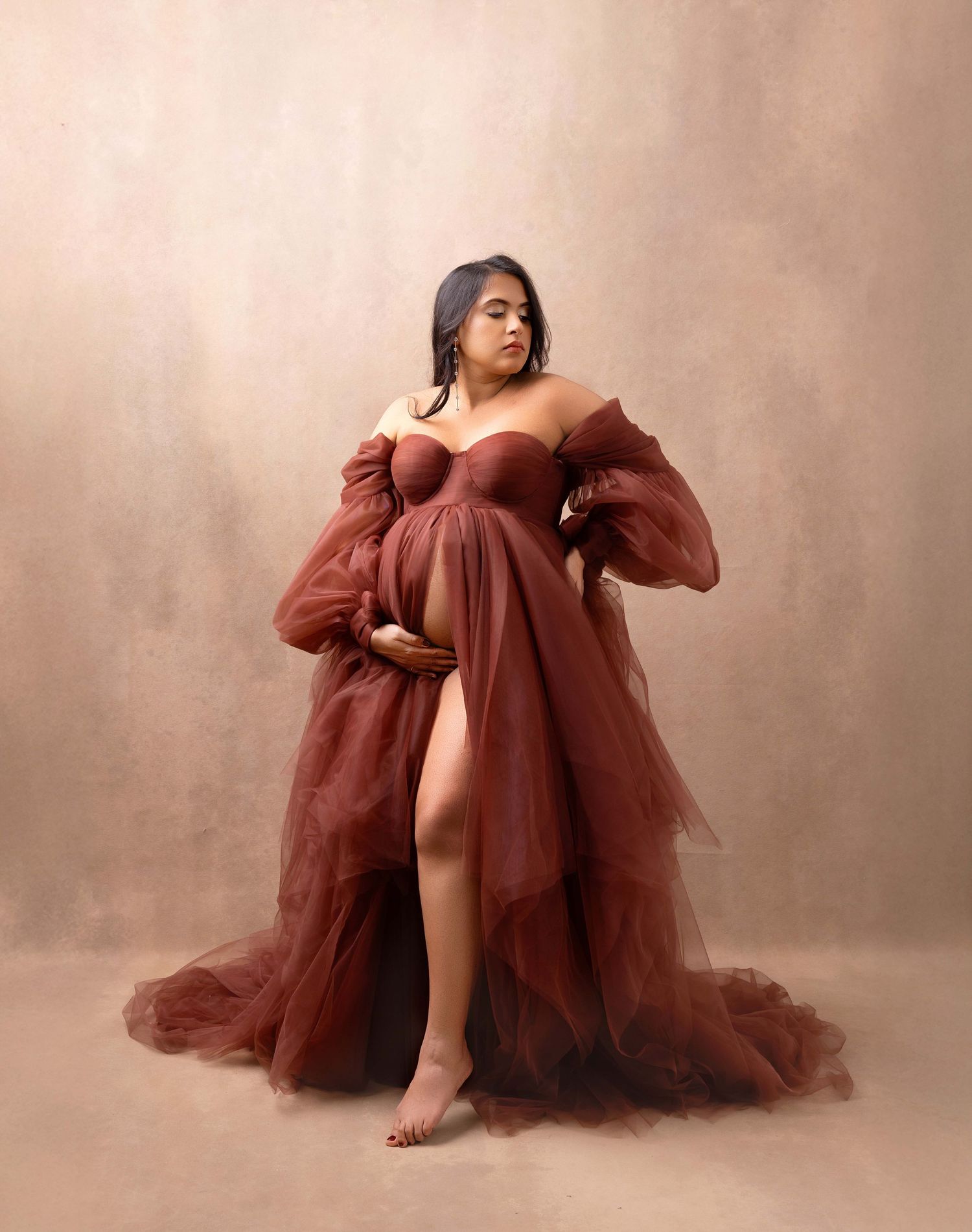 Ethereal maternity portrait in a flowing burgundy tulle gown against a soft beige backdrop in studio lighting.