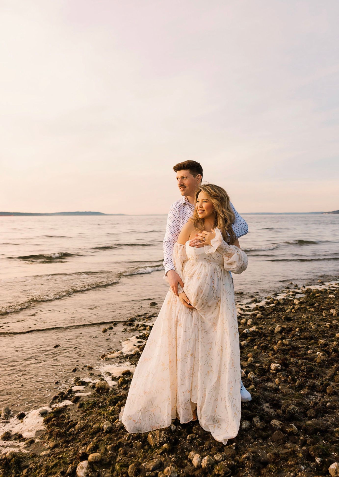 Couple embracing on rocky beach at sunset, woman in flowing white lace maternity gown, man in blue shirt, ocean backdrop.