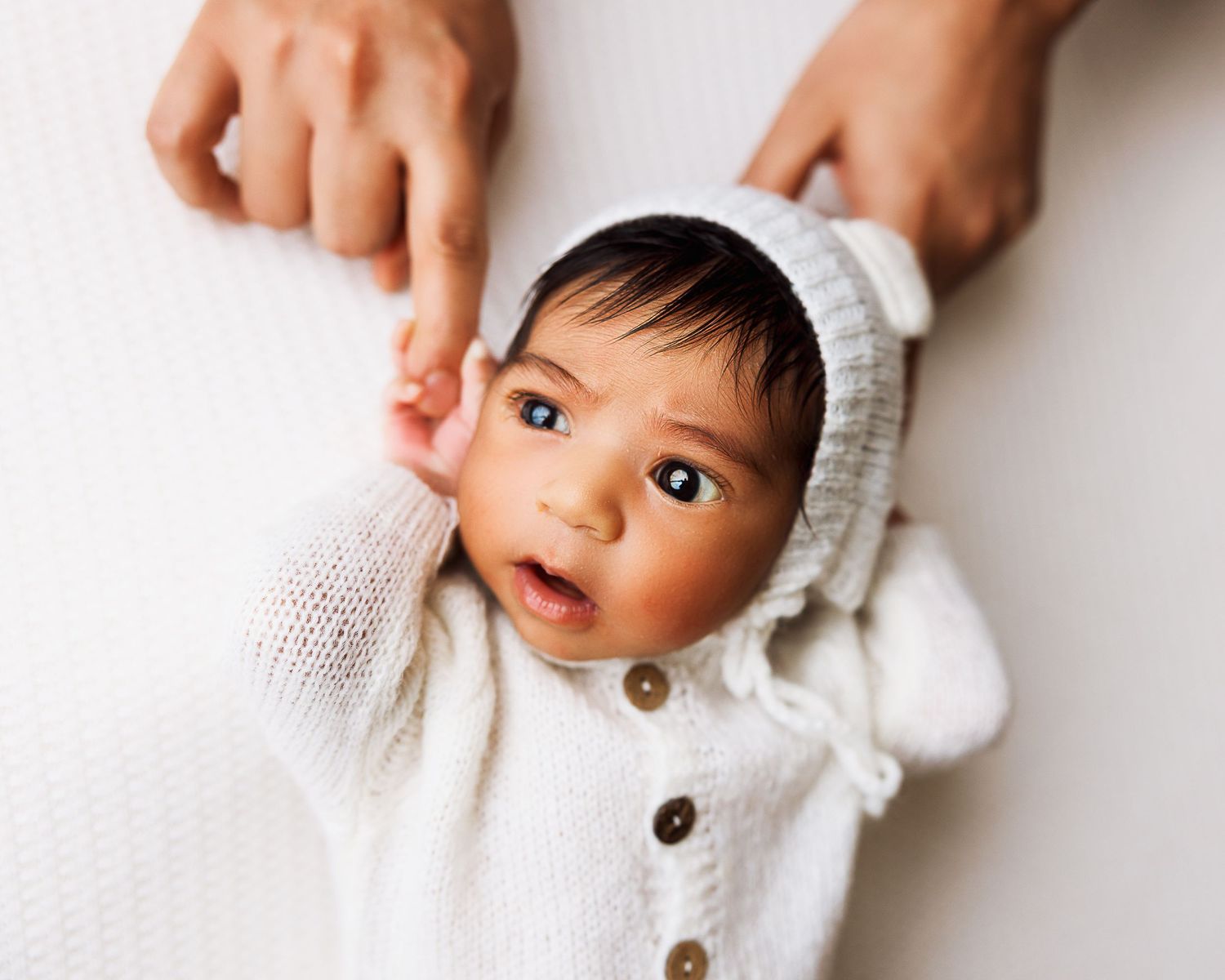 A baby wrapped in a cozy white knit blanket looks up with wide eyes while lying down.