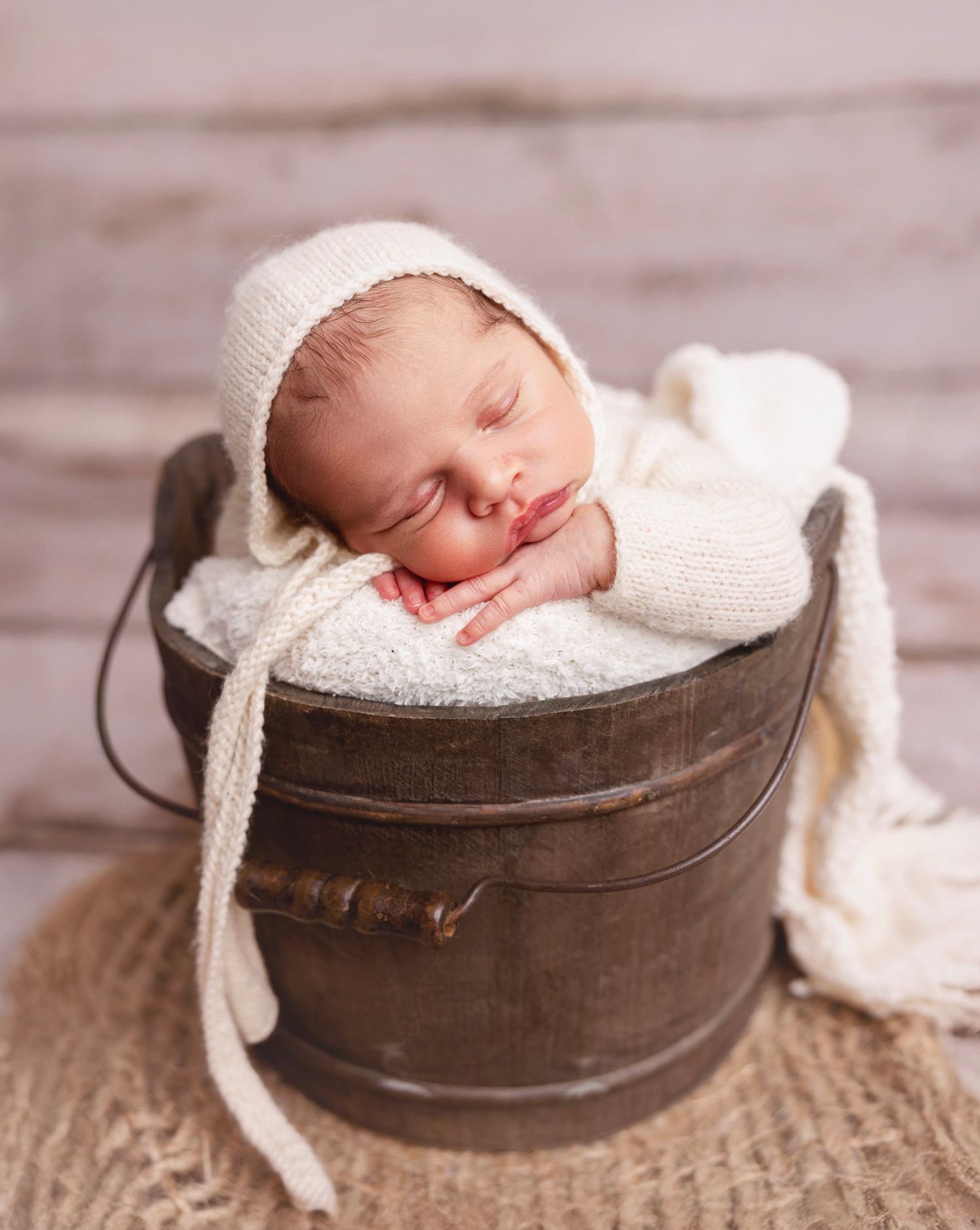 Sleeping newborn baby in cream knit bonnet nestled in rustic wooden bucket with white wrap blanket.