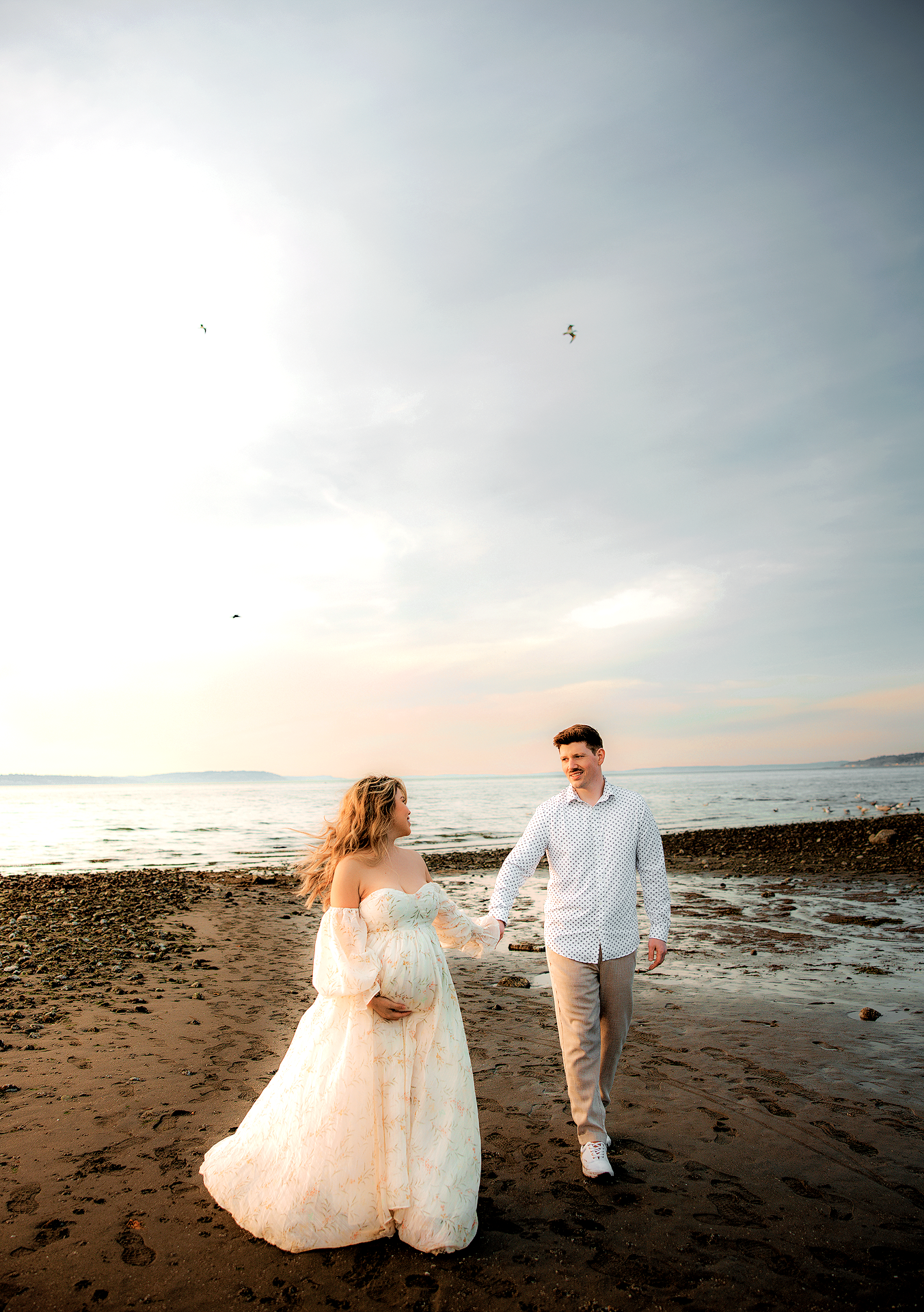 Pregnant couple holding hands on a rocky beach at sunset, woman in white lace maternity gown, man in white shirt.