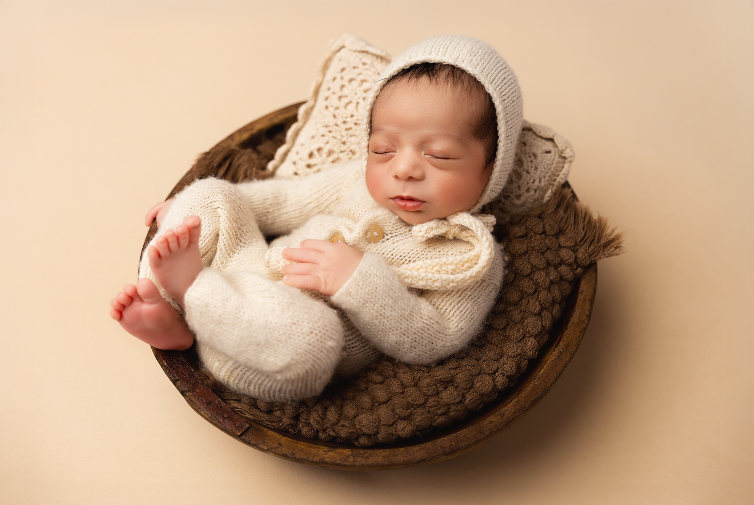 Sleeping newborn baby in cream knit bear romper and bonnet, nestled in a wooden bowl with brown wrap.