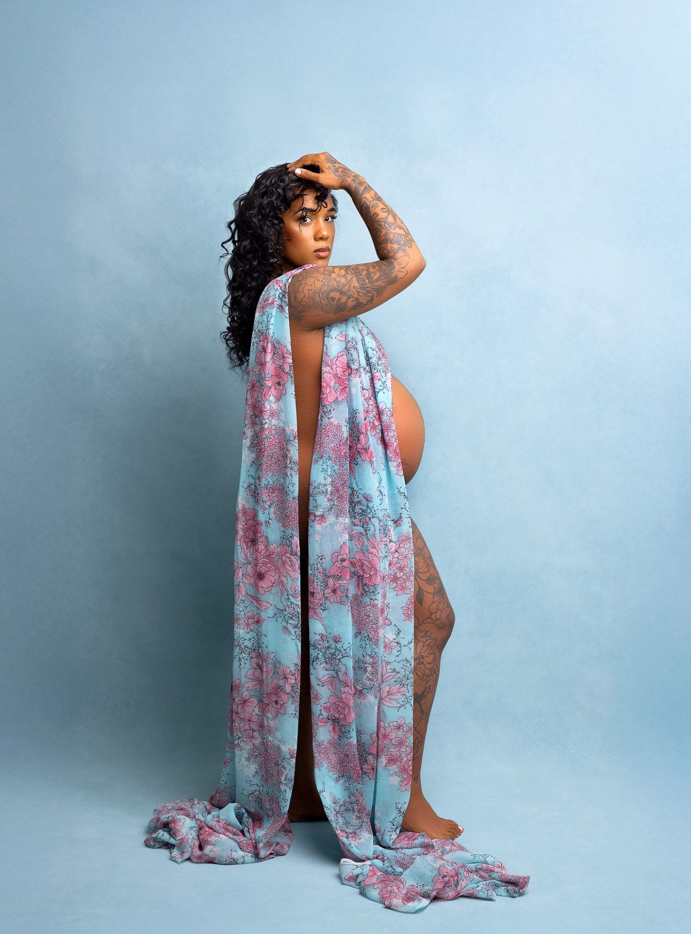 A figure in a flowing blue and pink floral dress poses artistically against a light blue studio backdrop.