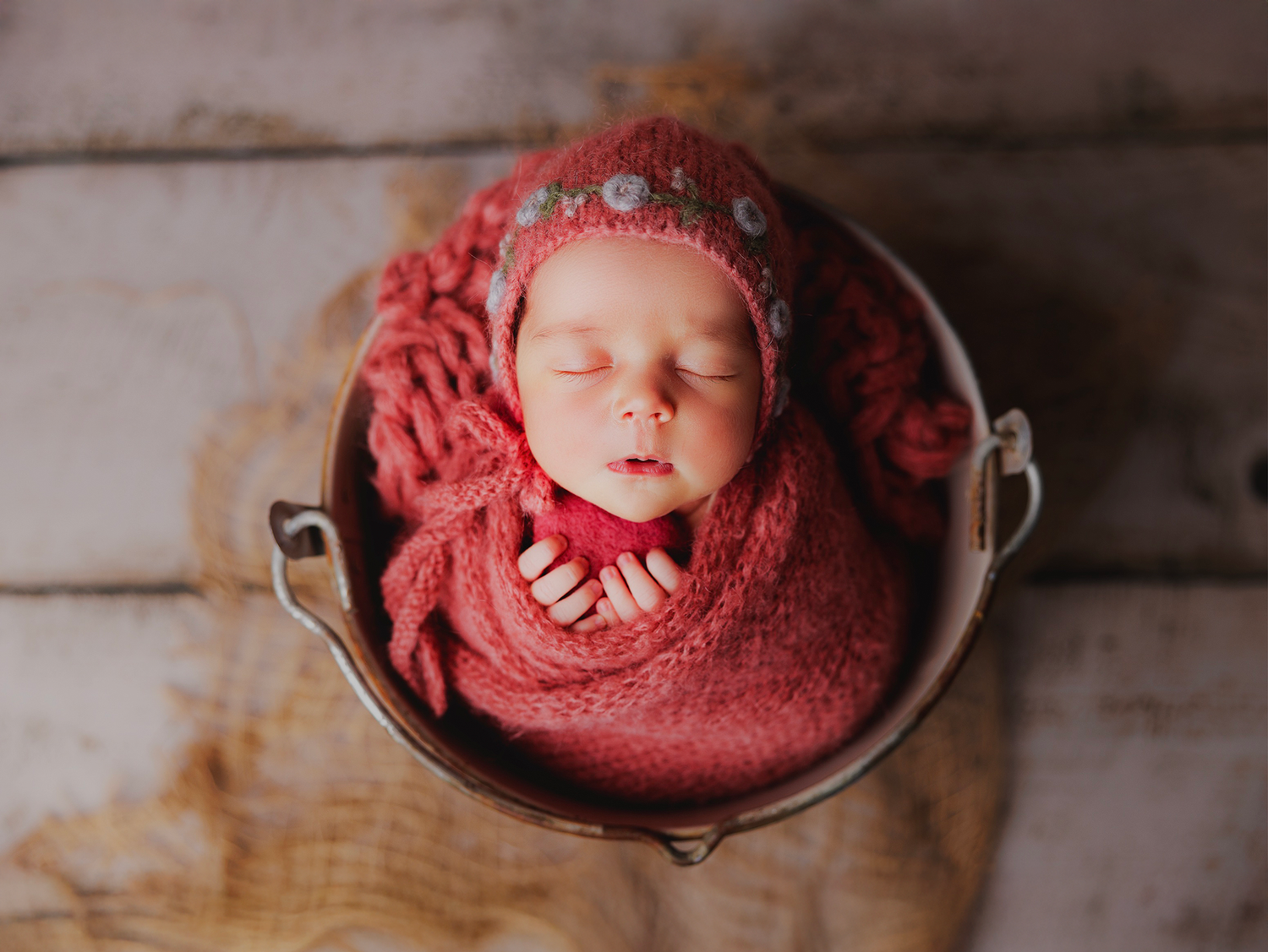 A sleeping newborn baby wrapped in red fabric lies peacefully in a rustic wooden bowl.