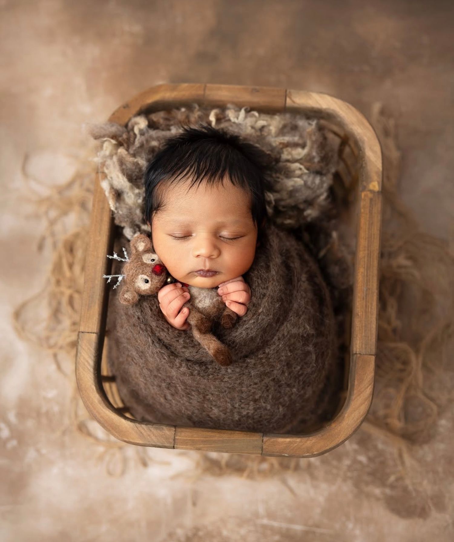 A sleeping newborn wrapped in a brown knit blanket rests peacefully in a wooden basket surrounded by soft feathers.