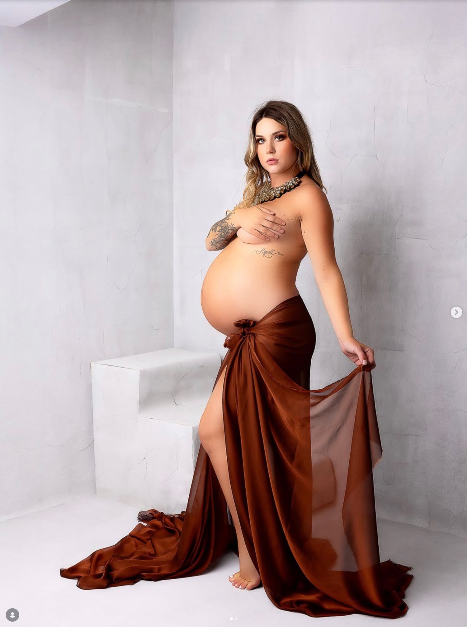 Artistic maternity photoshoot featuring a brown flowing dress against a minimalist white studio backdrop.